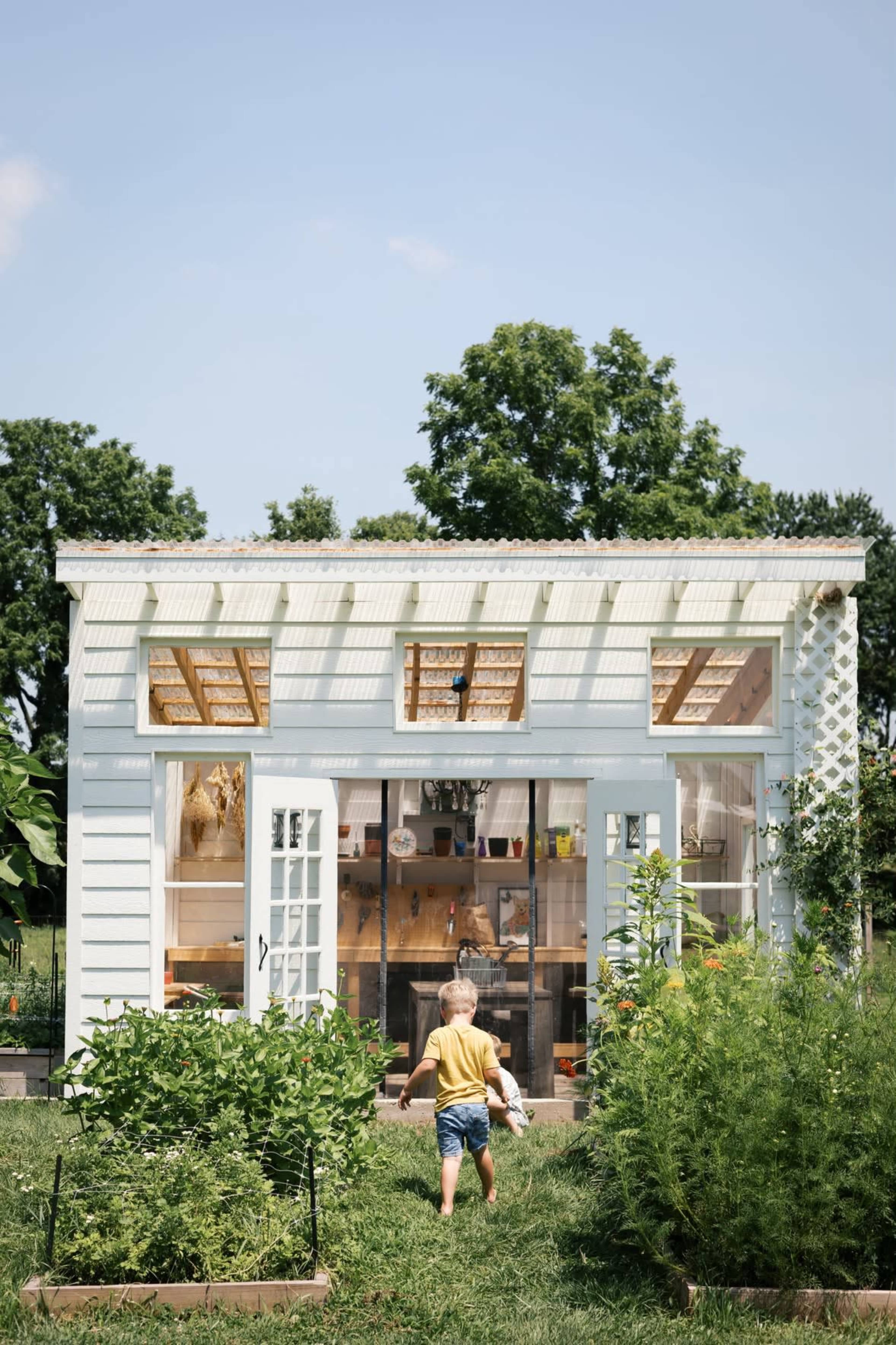 A child walks towards a small light blue shed surrounded by greenery and garden beds.