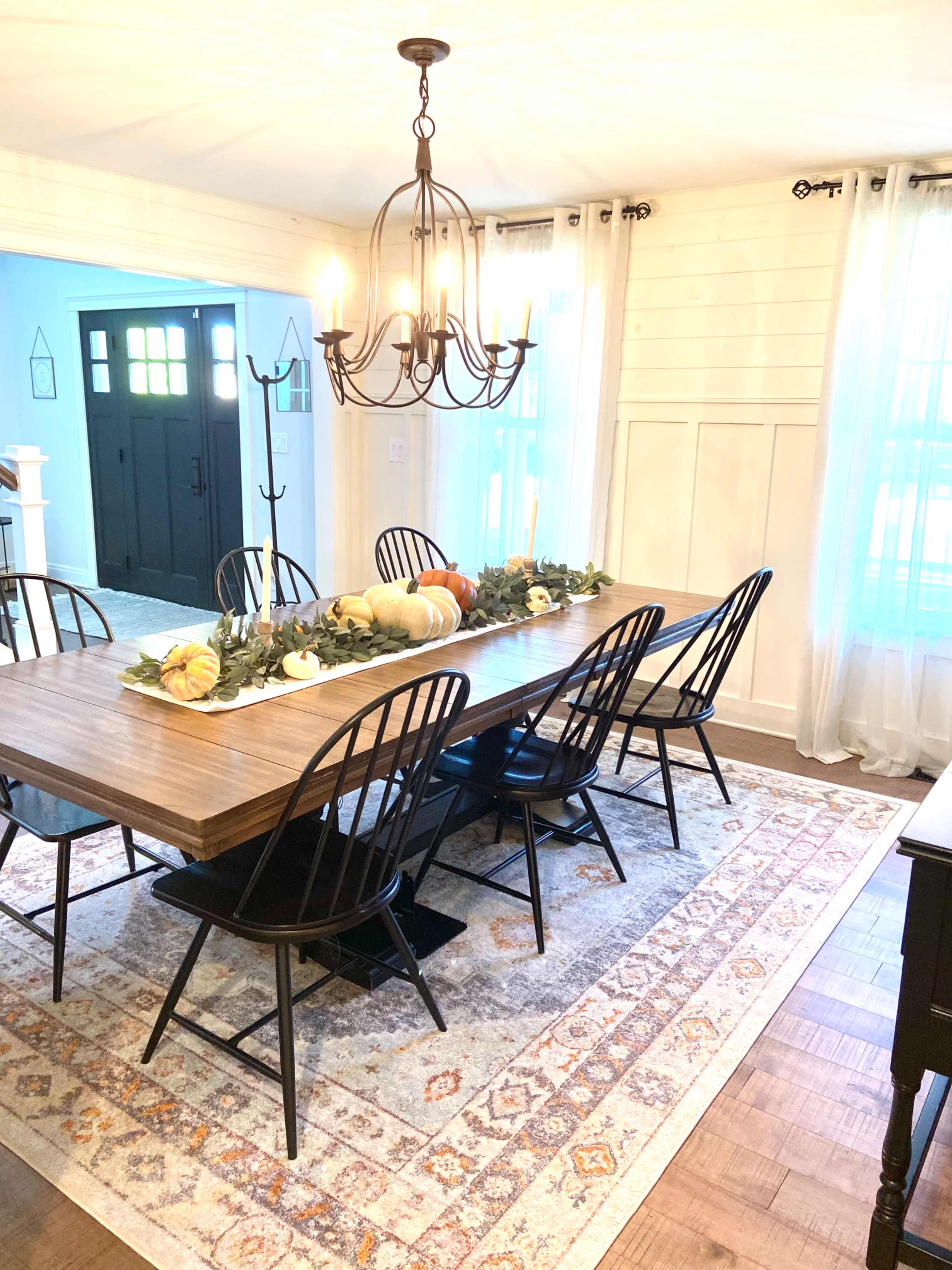 A dining room features a long wooden table with black chairs, adorned with a garland and decorative pumpkins, under a chandelier.