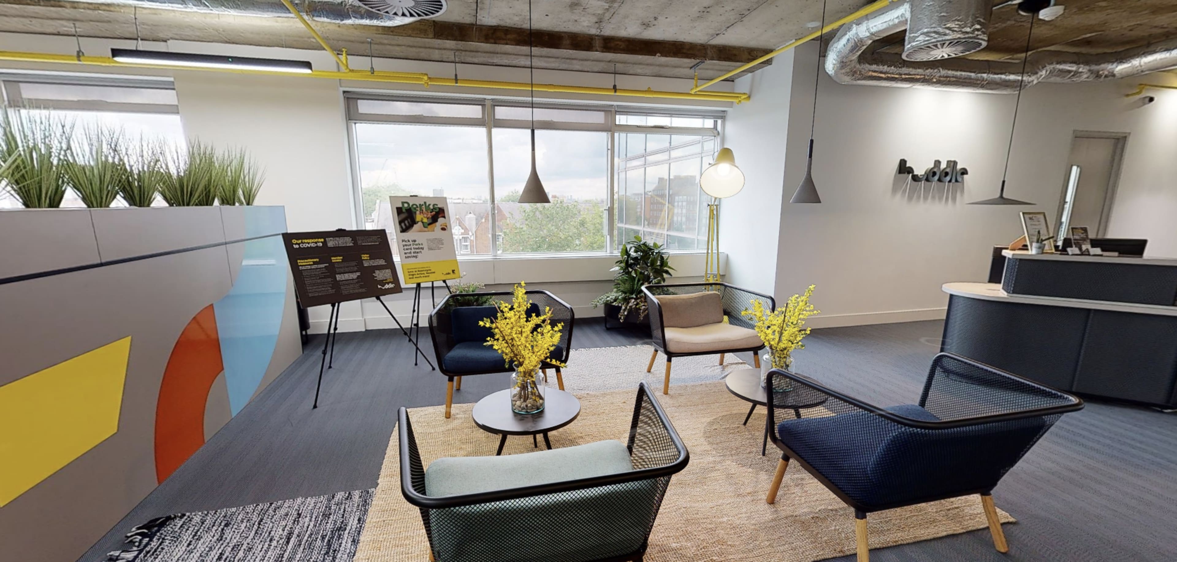 A modern office lounge area with several chairs, a small table, and plants, illuminated by natural light from large windows.