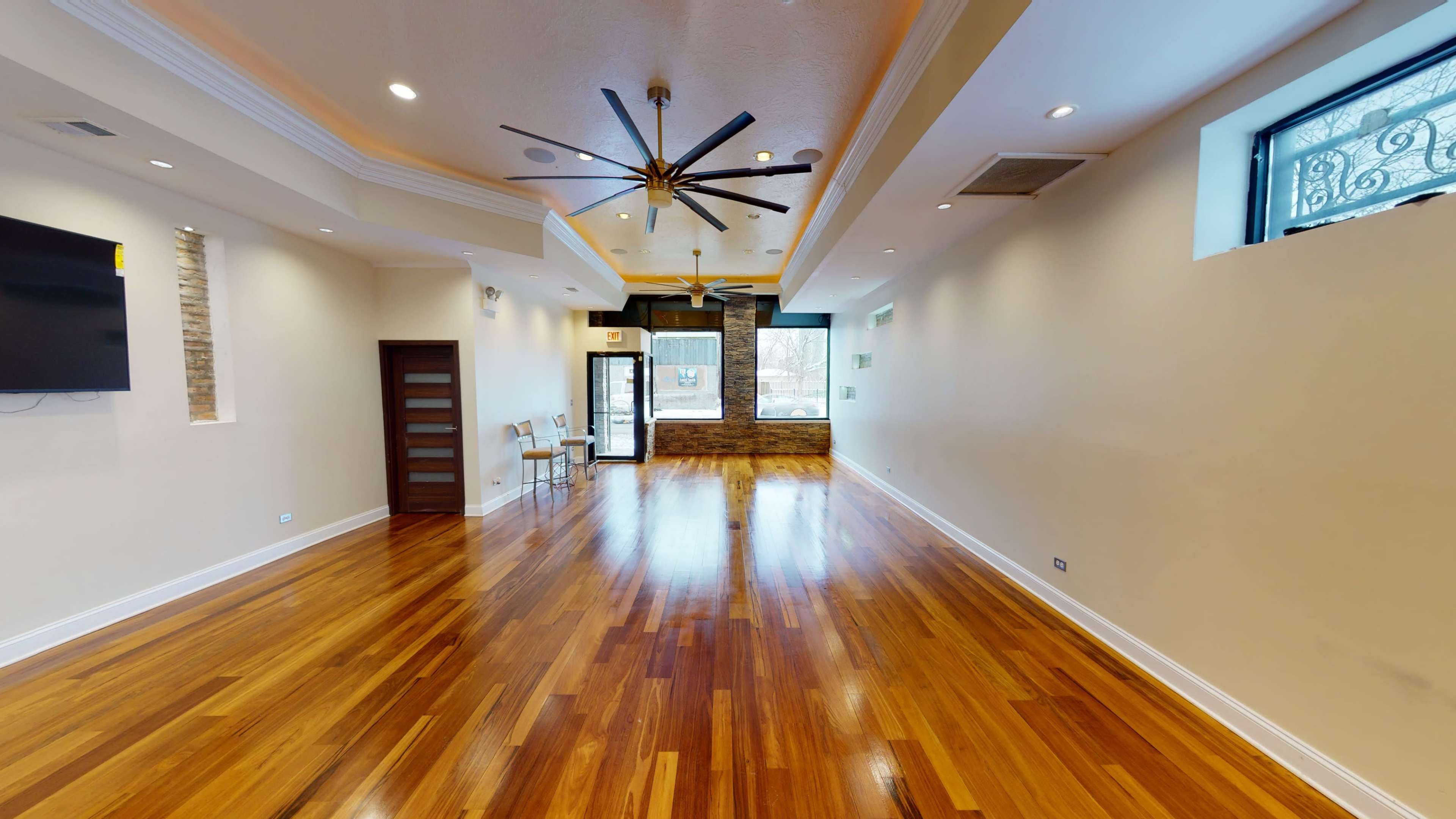 A spacious room with hardwood flooring, large ceiling fans, and natural light coming from windows at the far end.