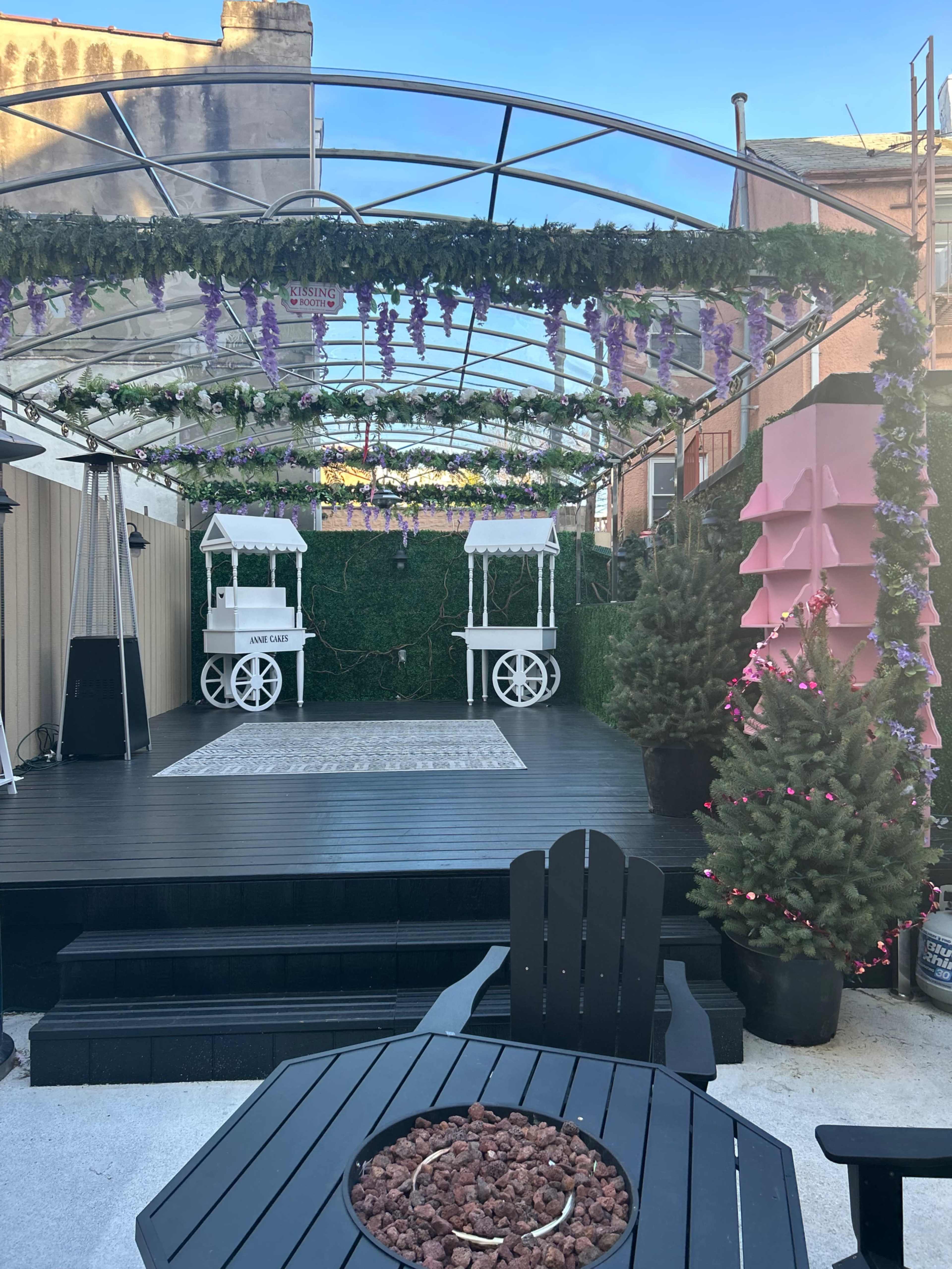 The scene features a garden area with a wooden deck, two white market-style stalls, a rug on the floor, and greenery adorned with purple plants and decorations.