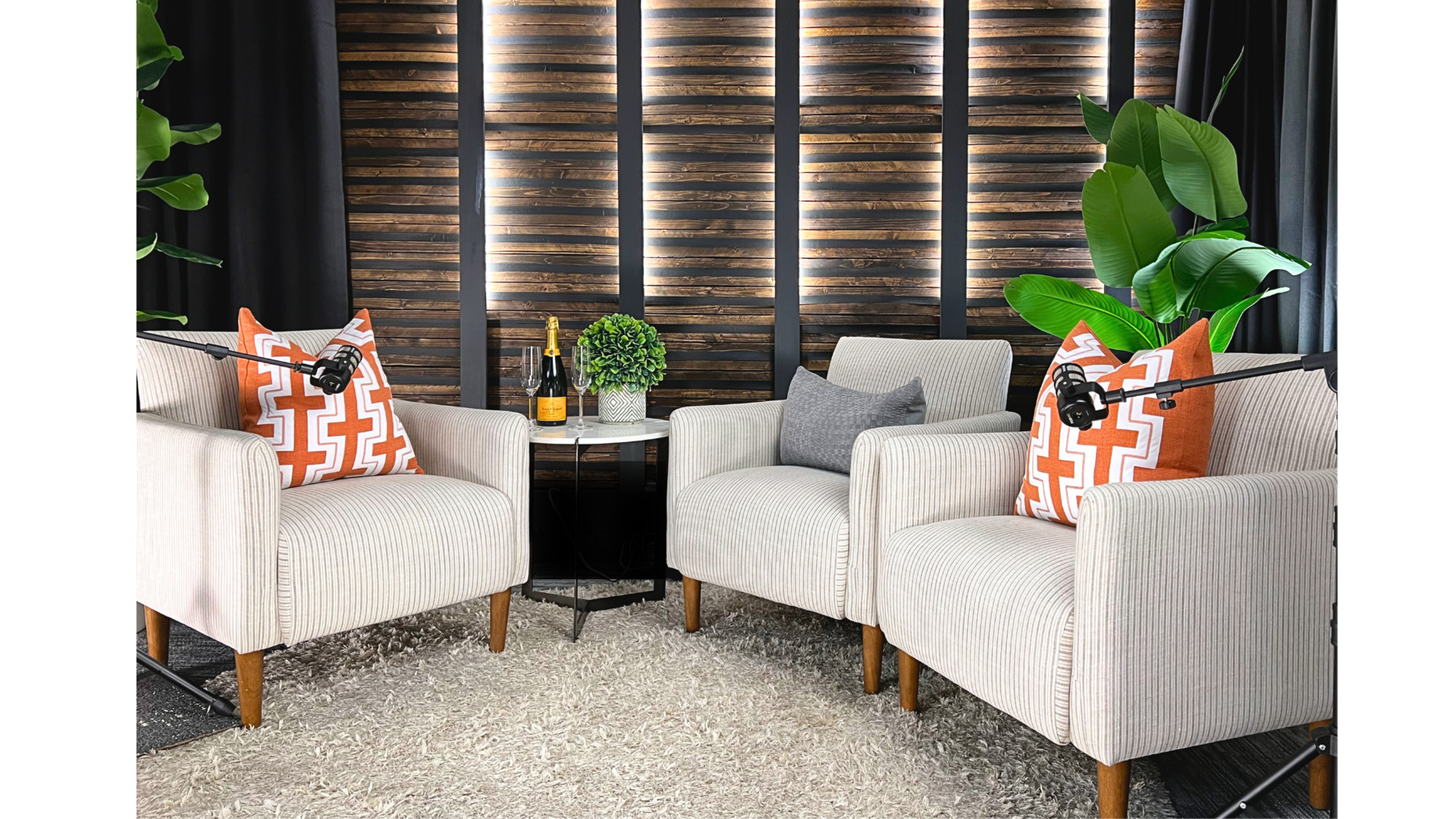 Two beige armchairs with patterned orange pillows are positioned around a small black table in a cozy setting featuring potted plants and a wooden paneled backdrop.
