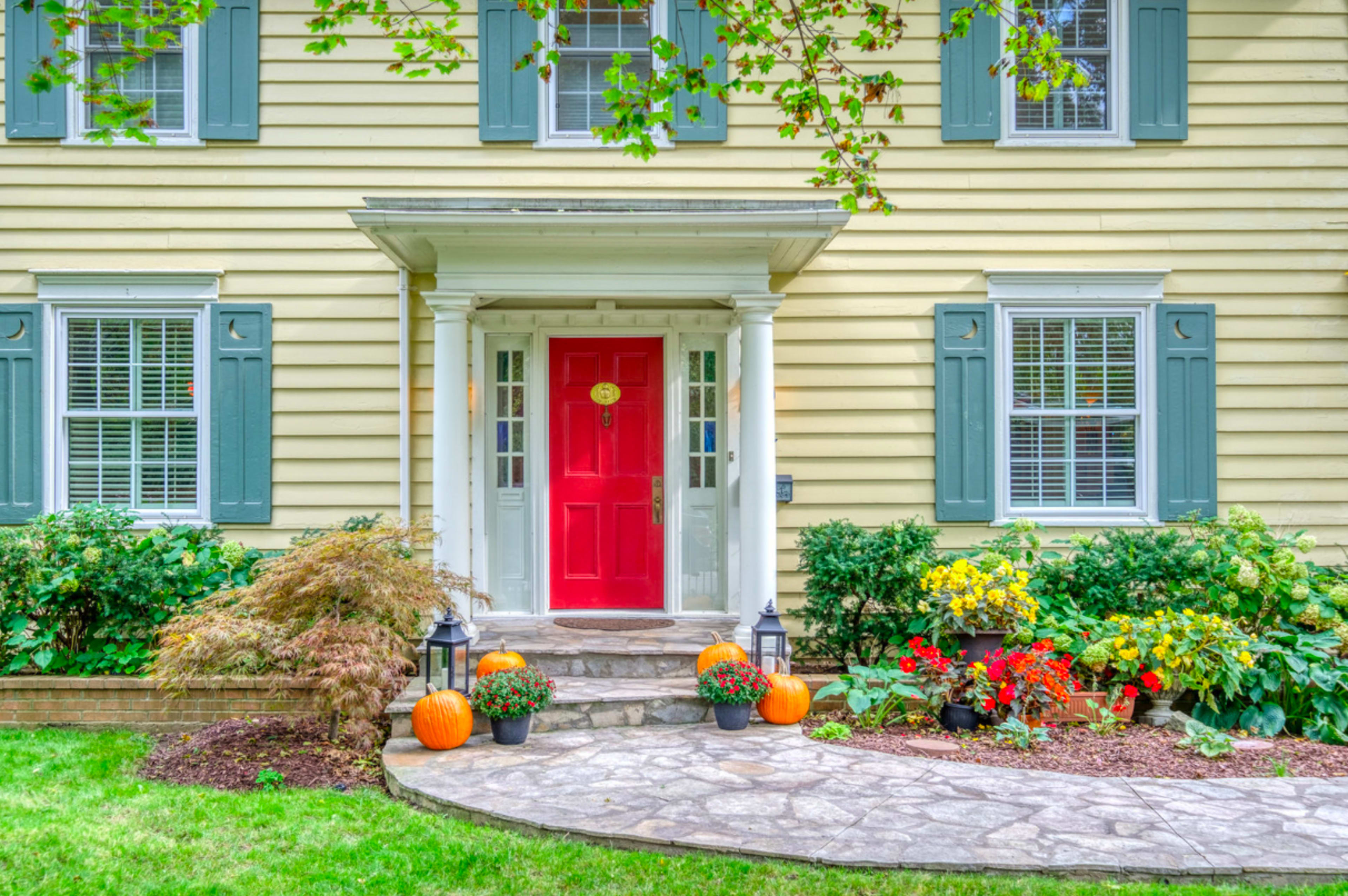 A yellow house with green shutters features a red door and a landscaped front yard adorned with pumpkins and colorful flowers.