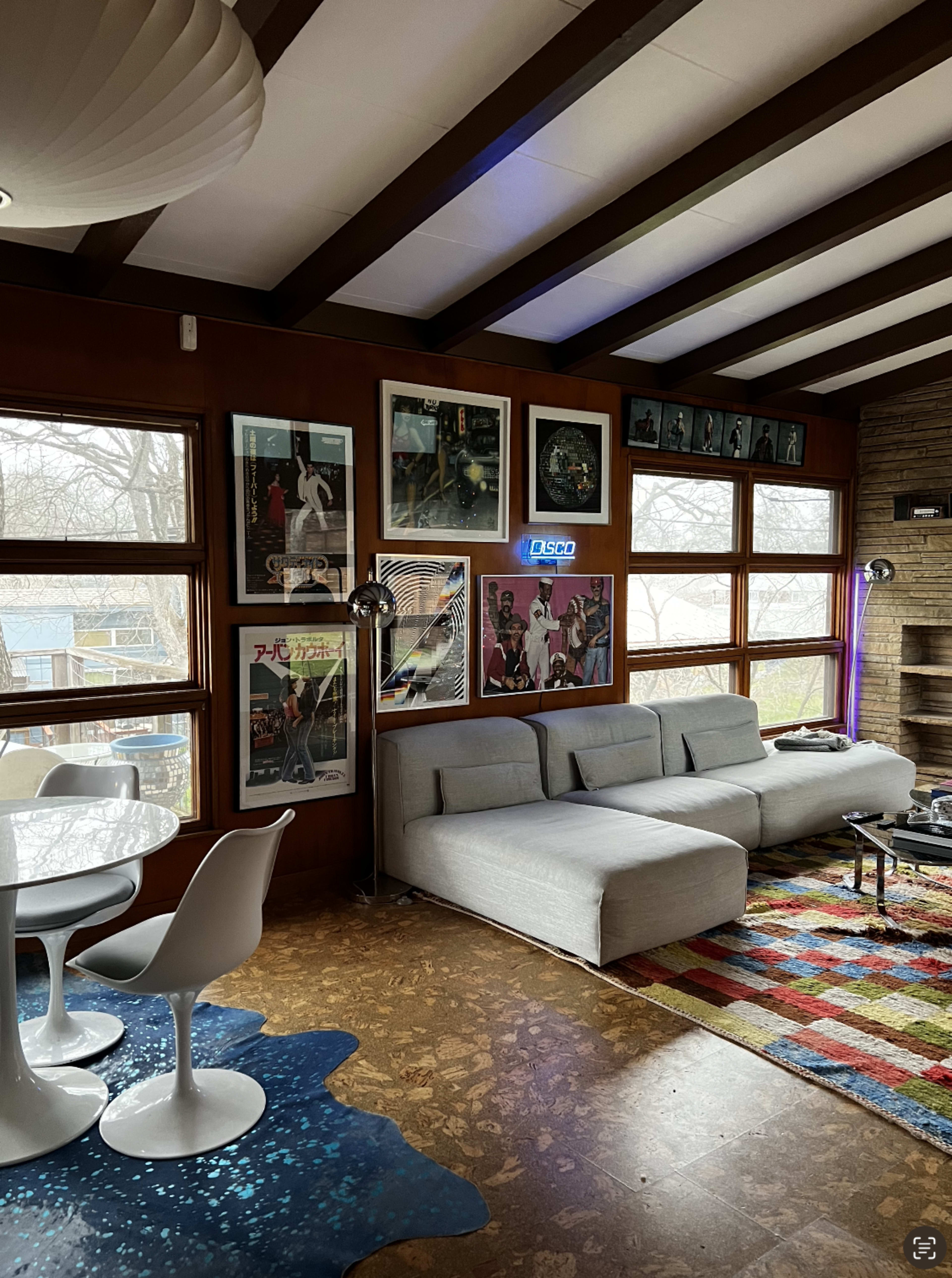 The image shows a modern living room with a gray sectional sofa, a round dining table with white chairs, and walls adorned with framed artwork and photographs.