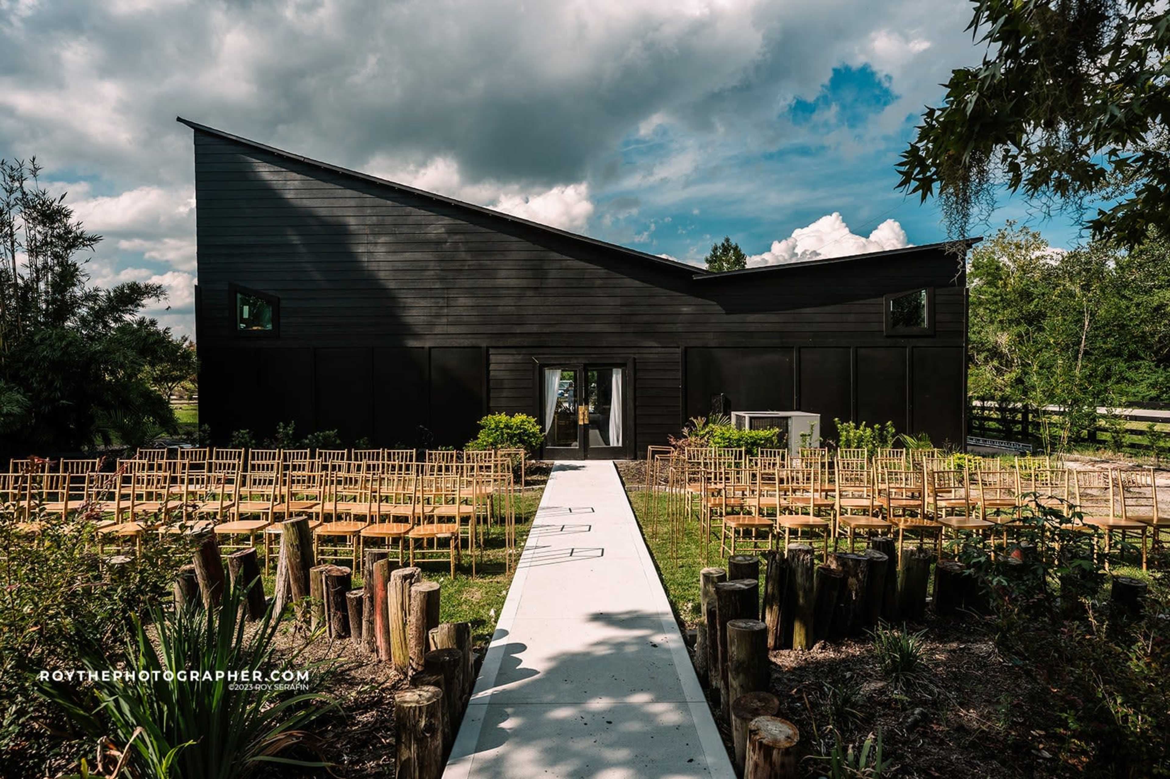 A modern black wooden building with a distinctive slanted roof, surrounded by rows of empty wooden chairs and a pathway lined with logs in a landscaped setting.