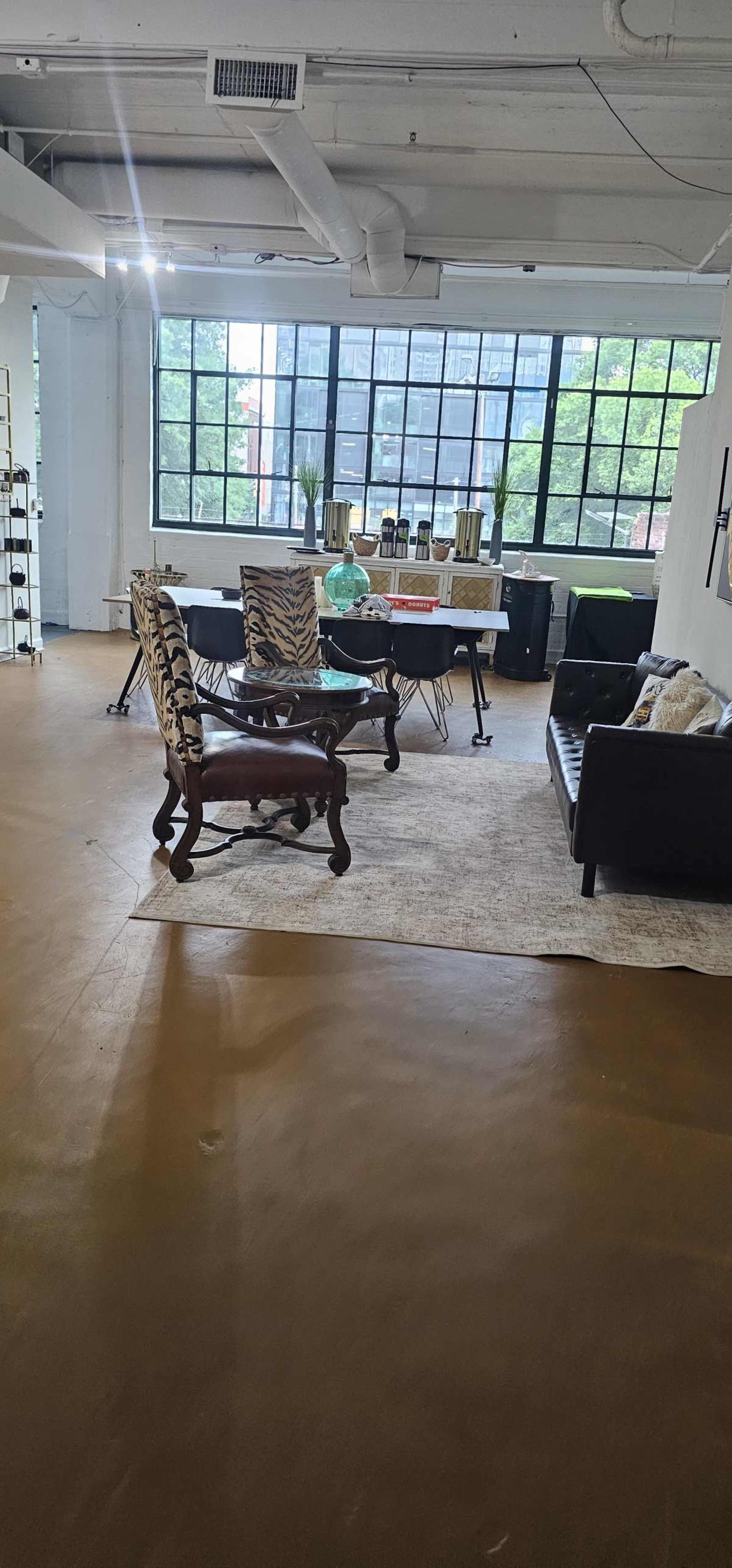 The image shows a spacious interior with a seating area featuring a dark leather sofa and two patterned chairs, a large window allowing natural light, and a table set with decorative items.