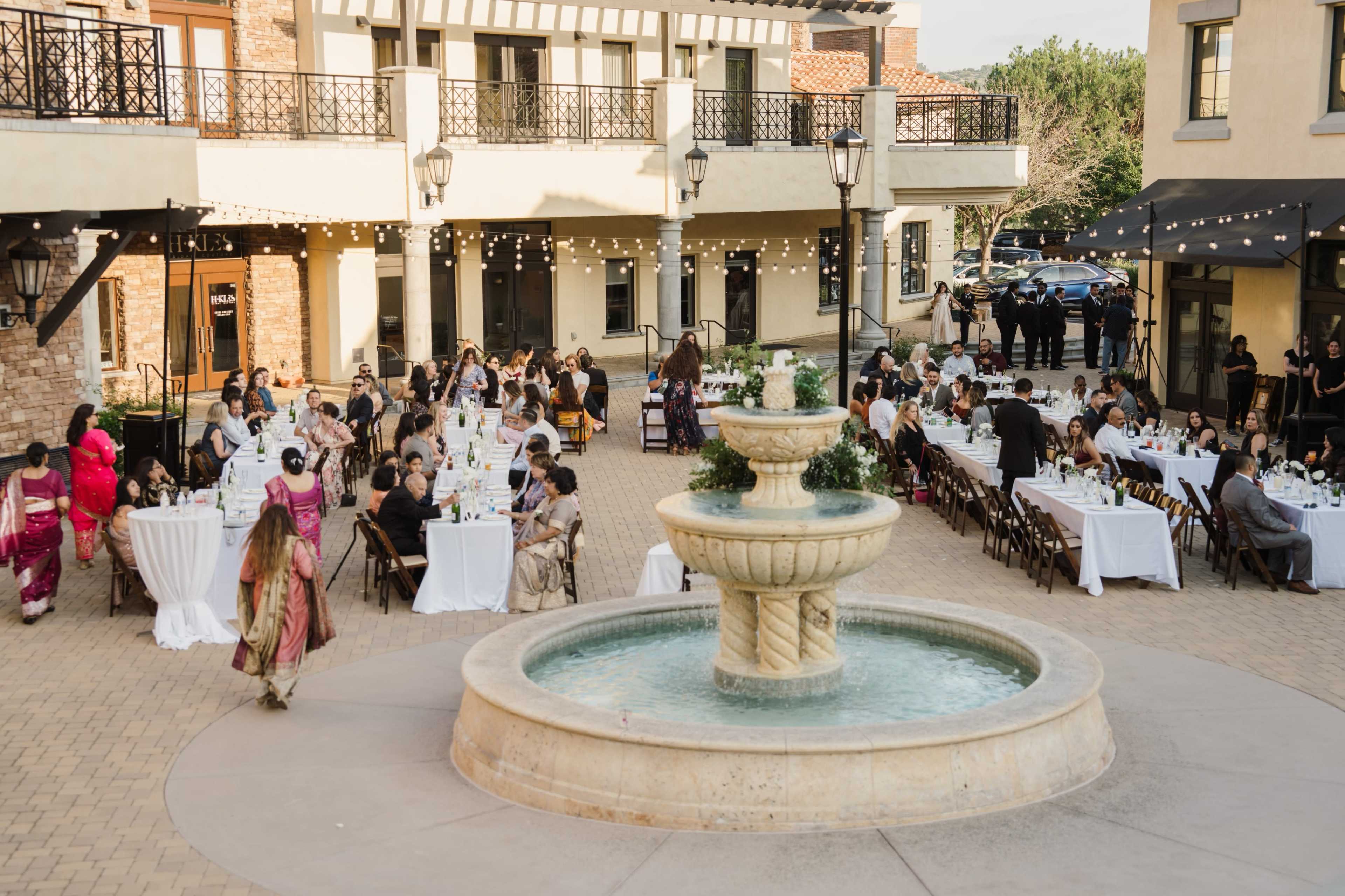A large outdoor gathering with guests seated at tables surrounding a central fountain.