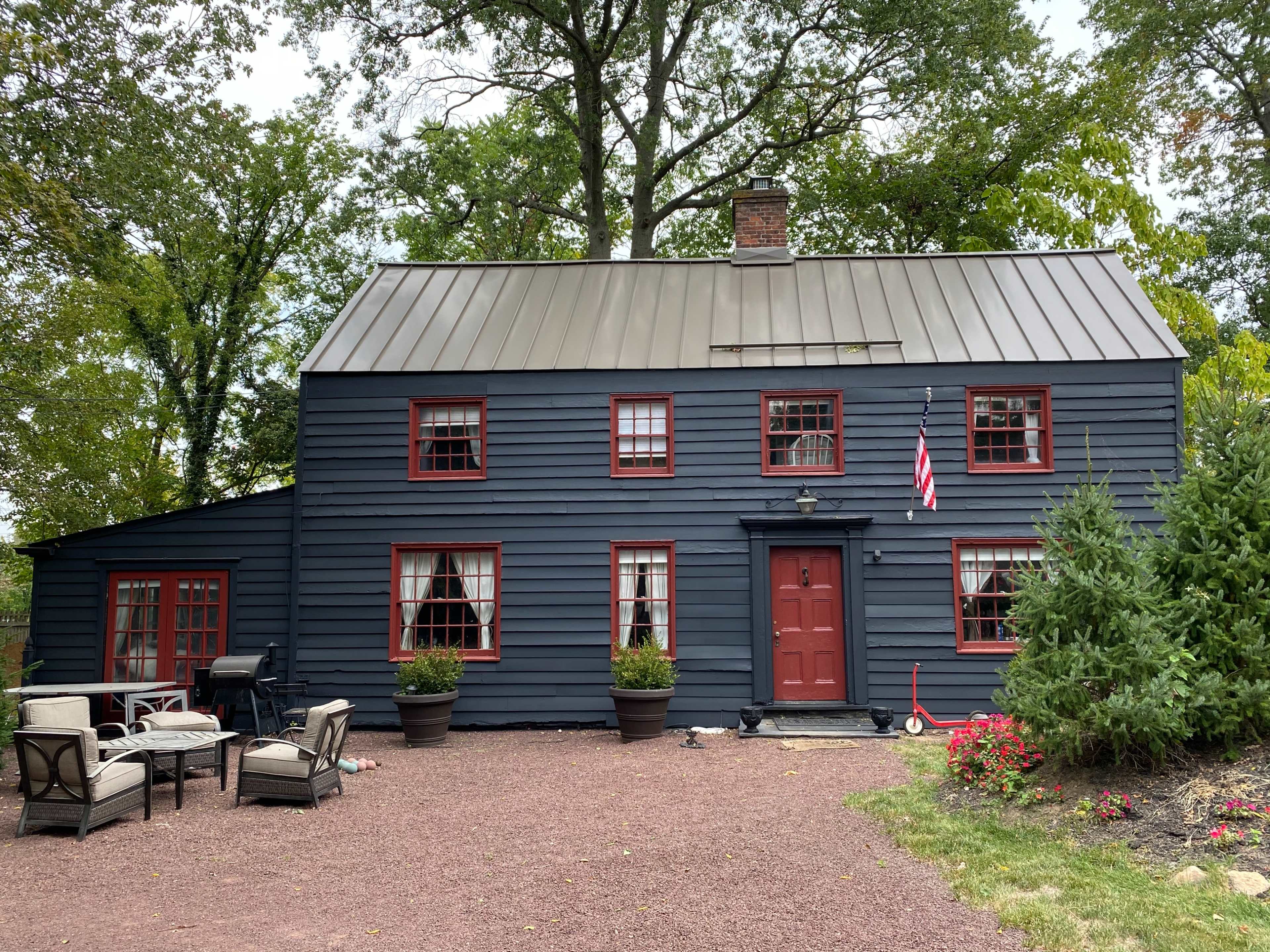 The image shows a dark blue wooden house with red trim and multiple windows, accompanied by outdoor seating arrangements and garden plants in front.