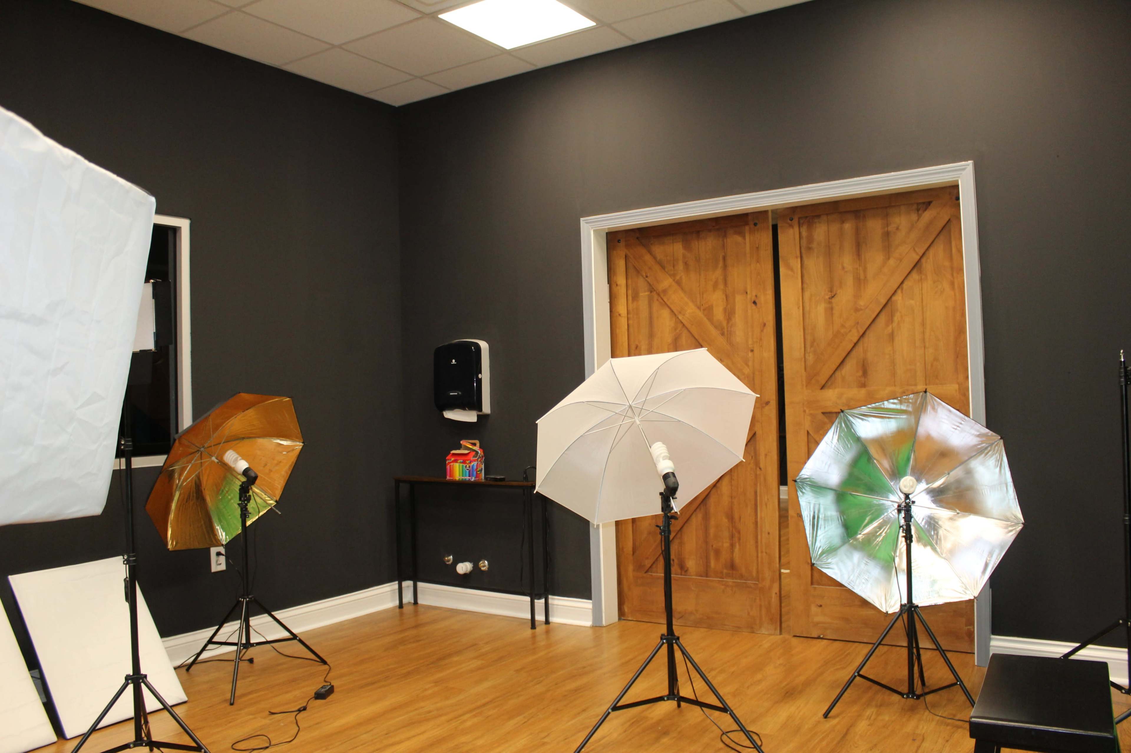 The image shows a photography studio setup with three lighting umbrellas, a wooden door, and a black wall.