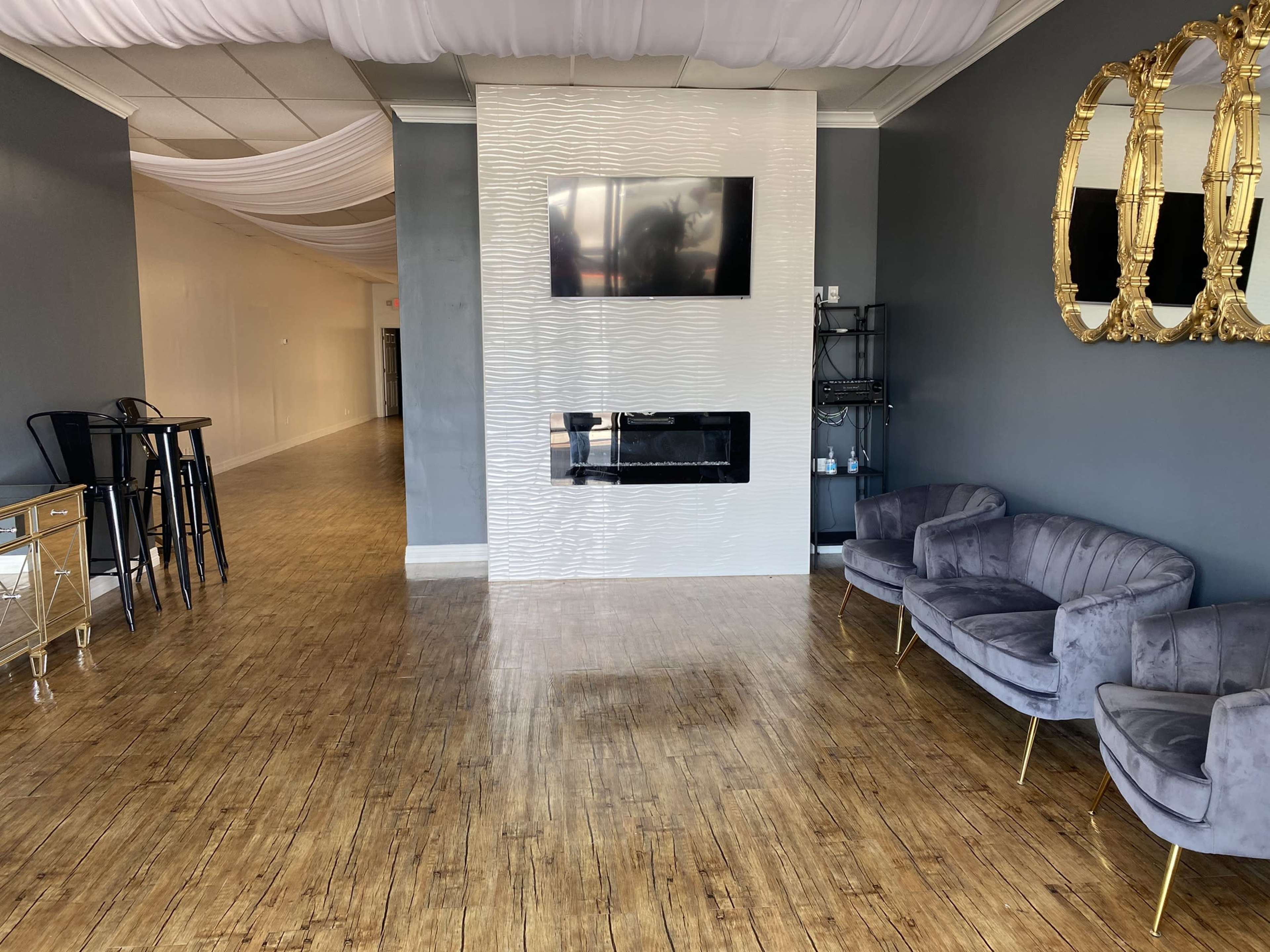 The image shows a spacious interior room with a modern electric fireplace, a wall-mounted TV, elegant gold-framed mirrors, plush chairs, and a sleek bar table.