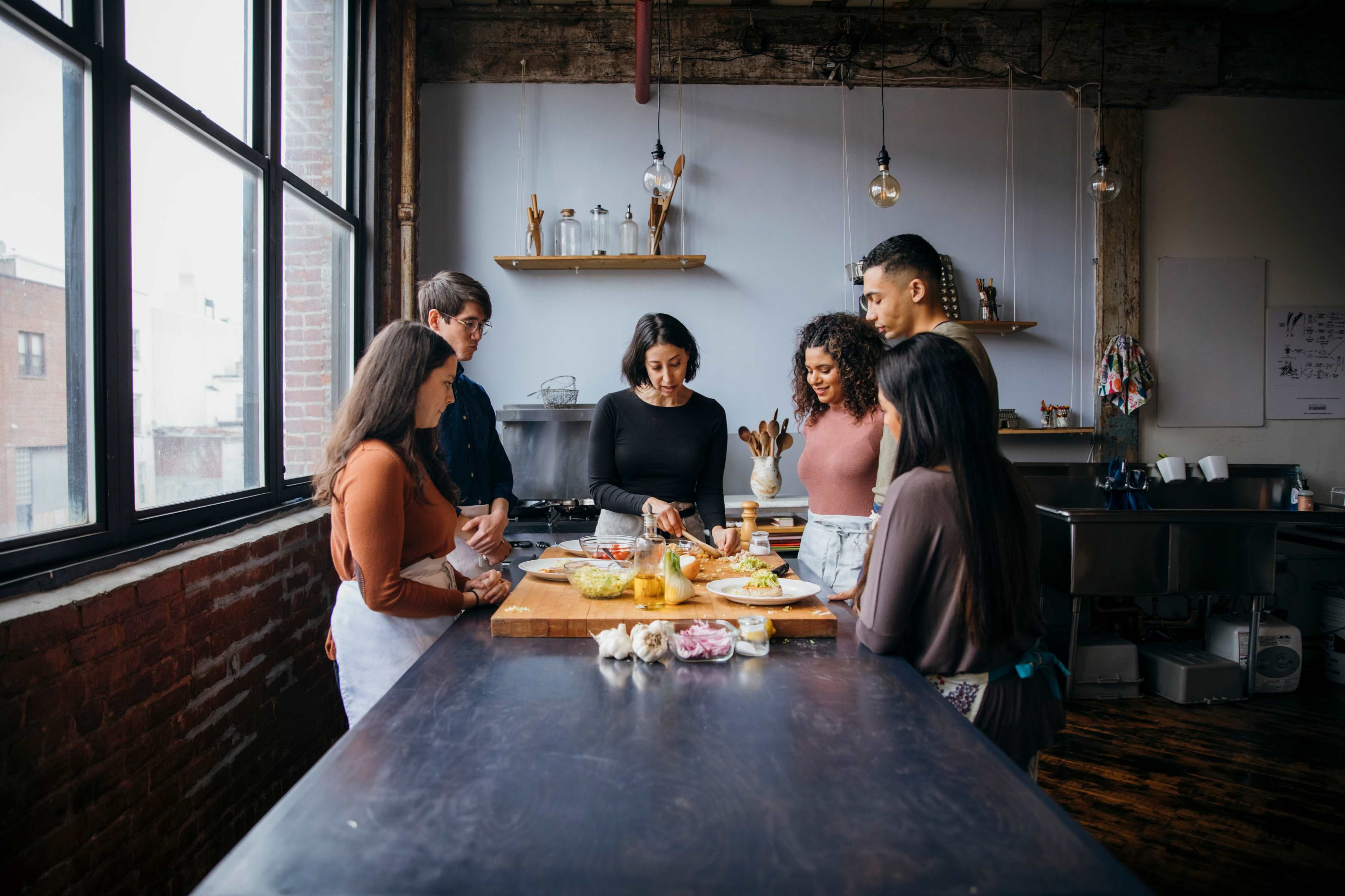 A group of six people is gathered around a kitchen island, preparing food together in a well-lit kitchen.