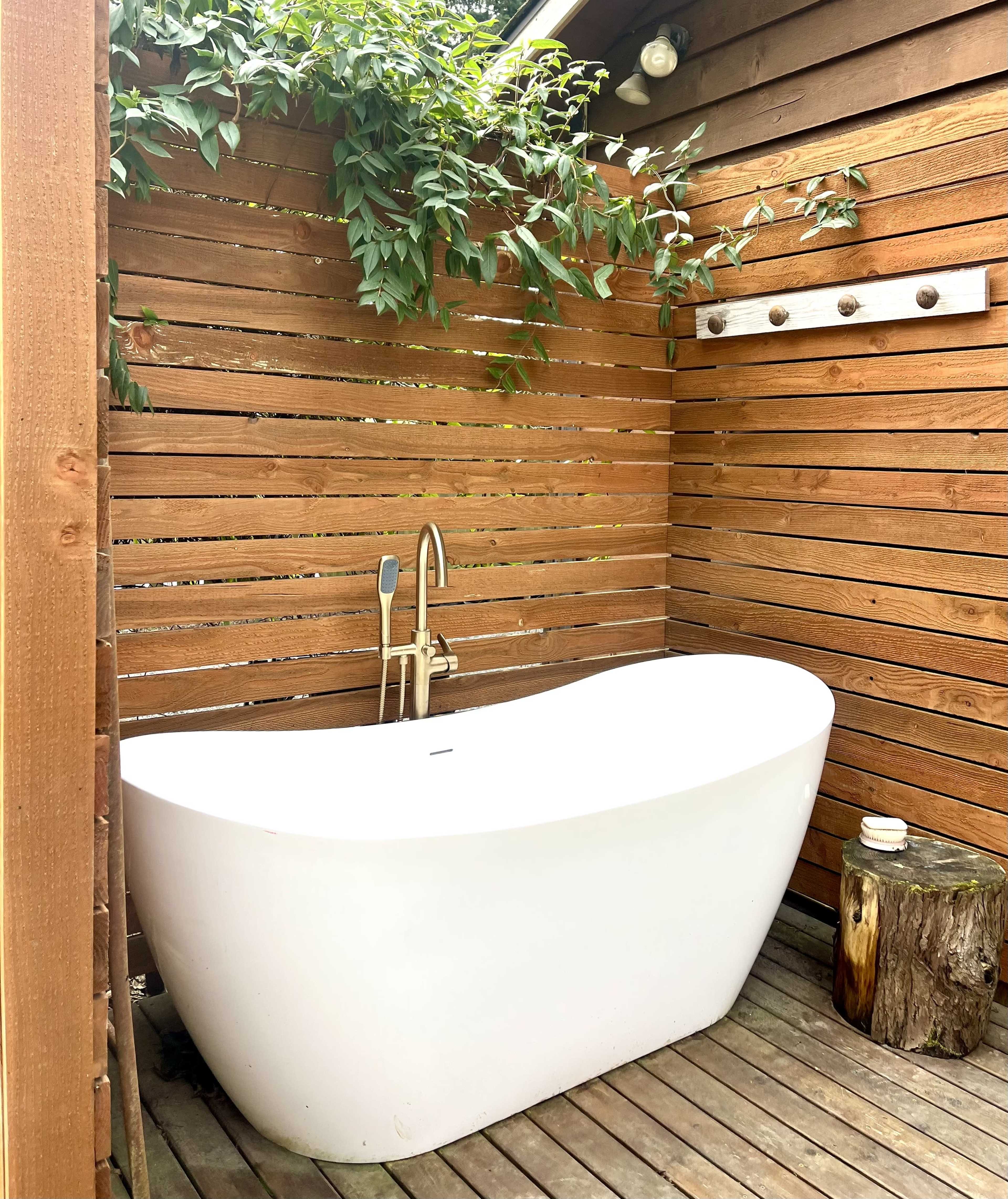 The image shows an outdoor bathroom featuring a freestanding white bathtub against wooden slatted walls, surrounded by greenery.