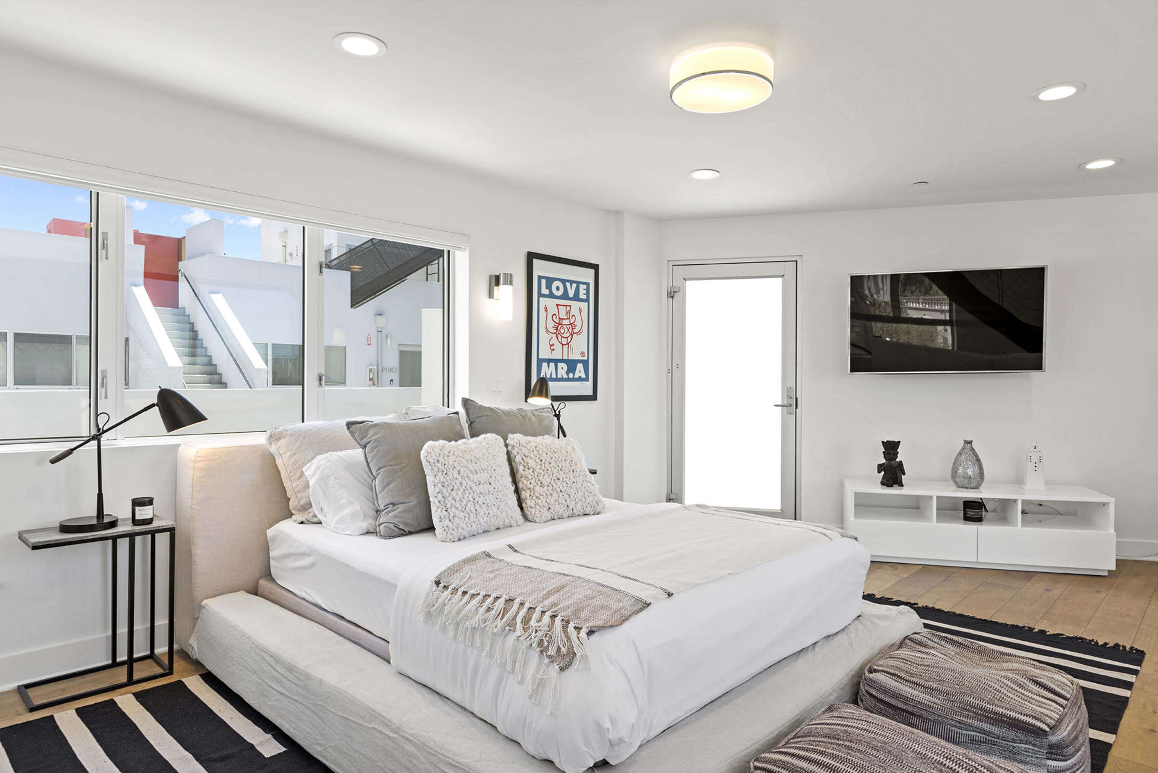 A modern bedroom featuring a low platform bed with gray bedding, a bedside lamp, a minimalist white entertainment unit, and large windows letting in natural light.