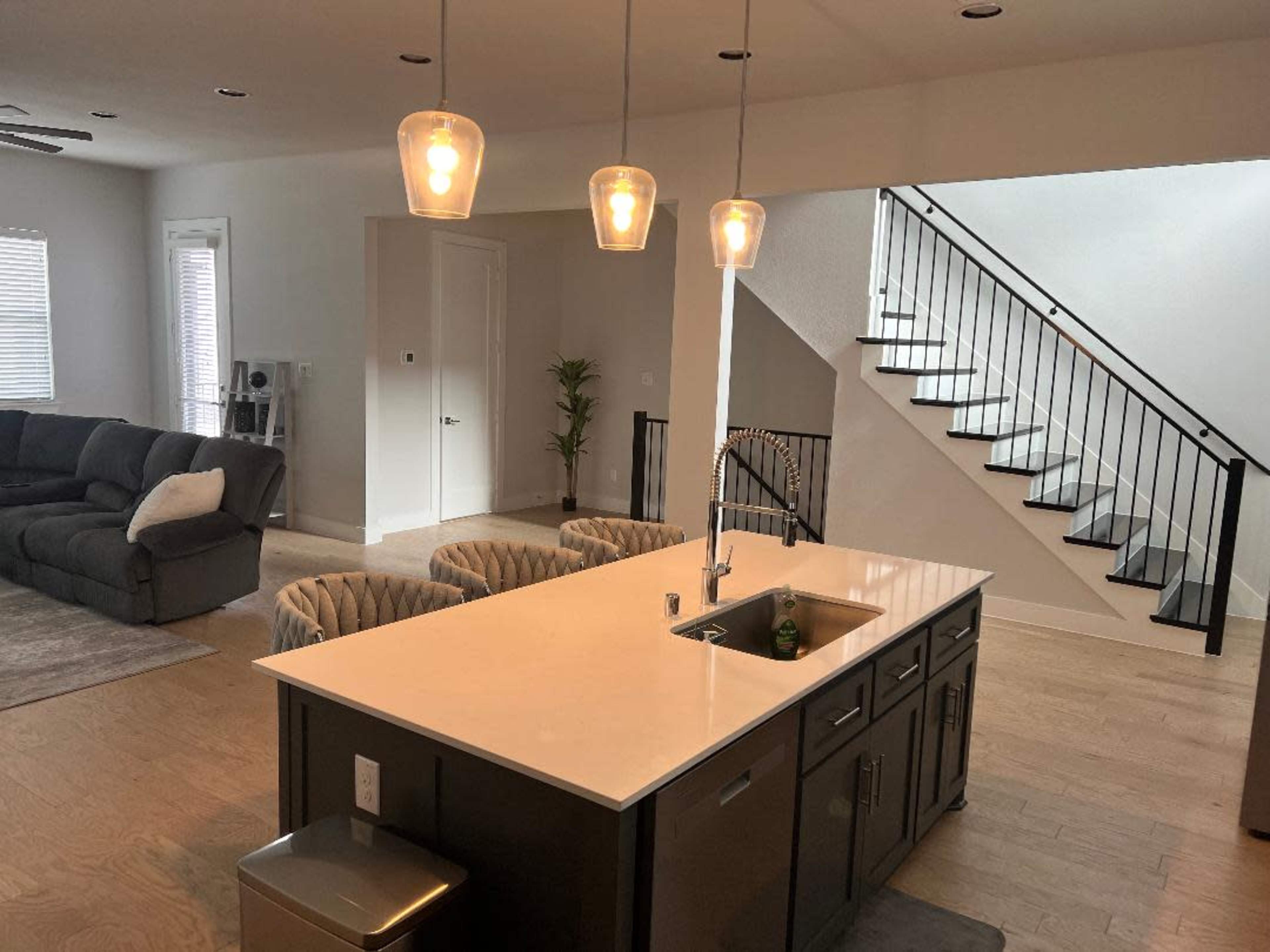 A modern kitchen with a large island, three pendant lights above it, and a staircase in the background leading to an upper level.