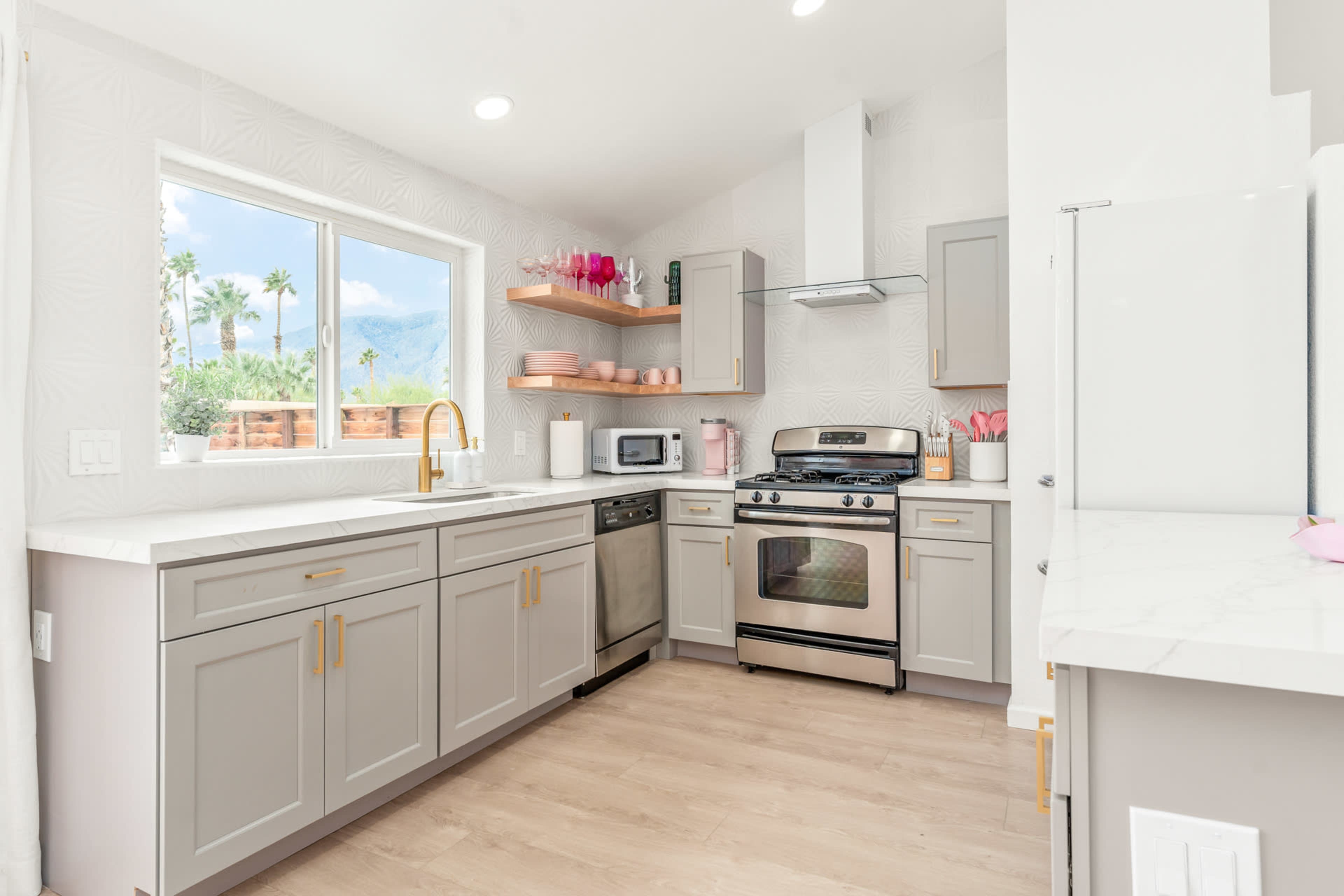 A modern kitchen featuring light gray cabinets, stainless steel appliances, and a window with a view of palm trees outside.