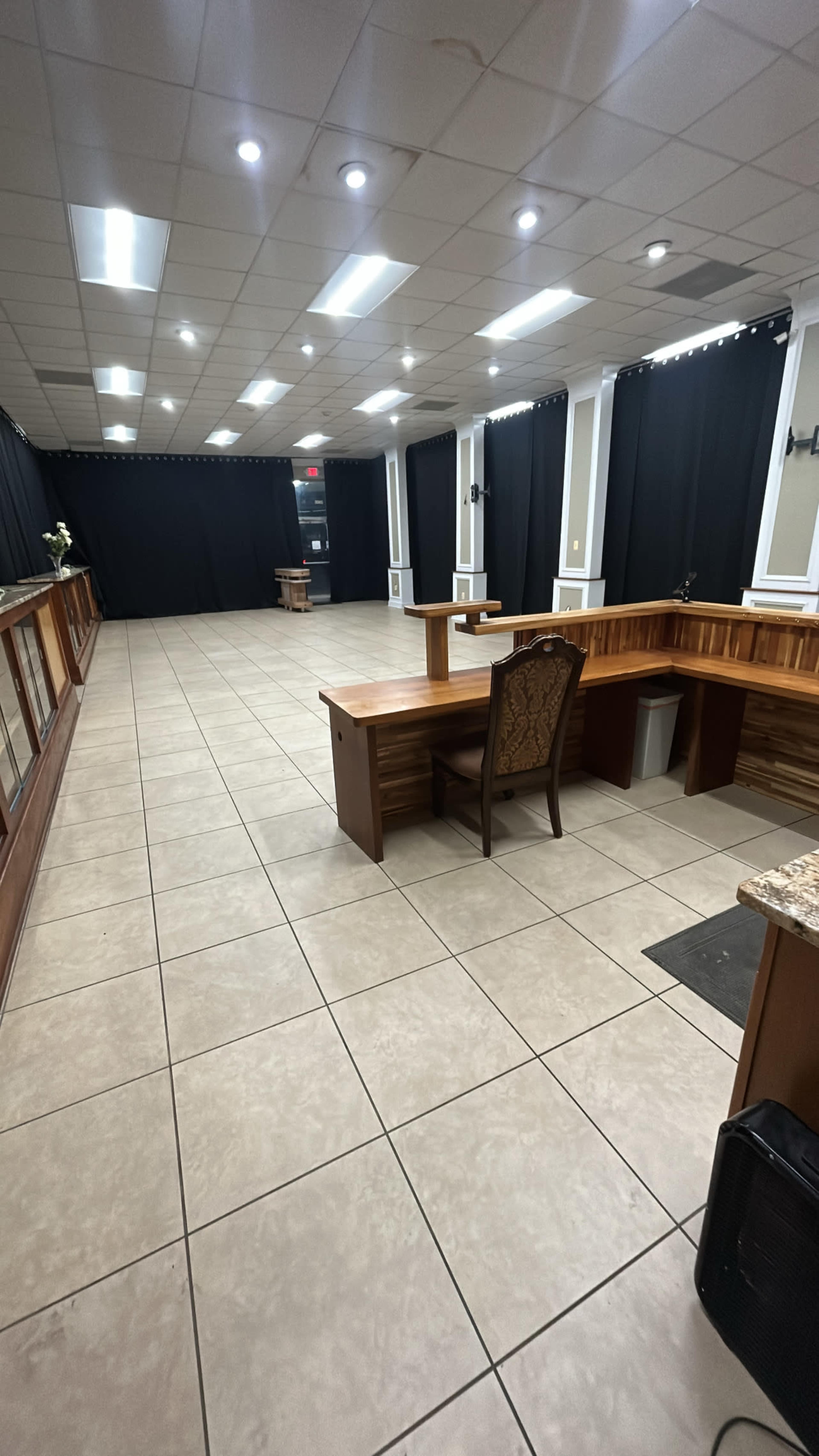 An empty room with tiled flooring, featuring wooden counters, a desk with a chair, and black curtains covering the windows.