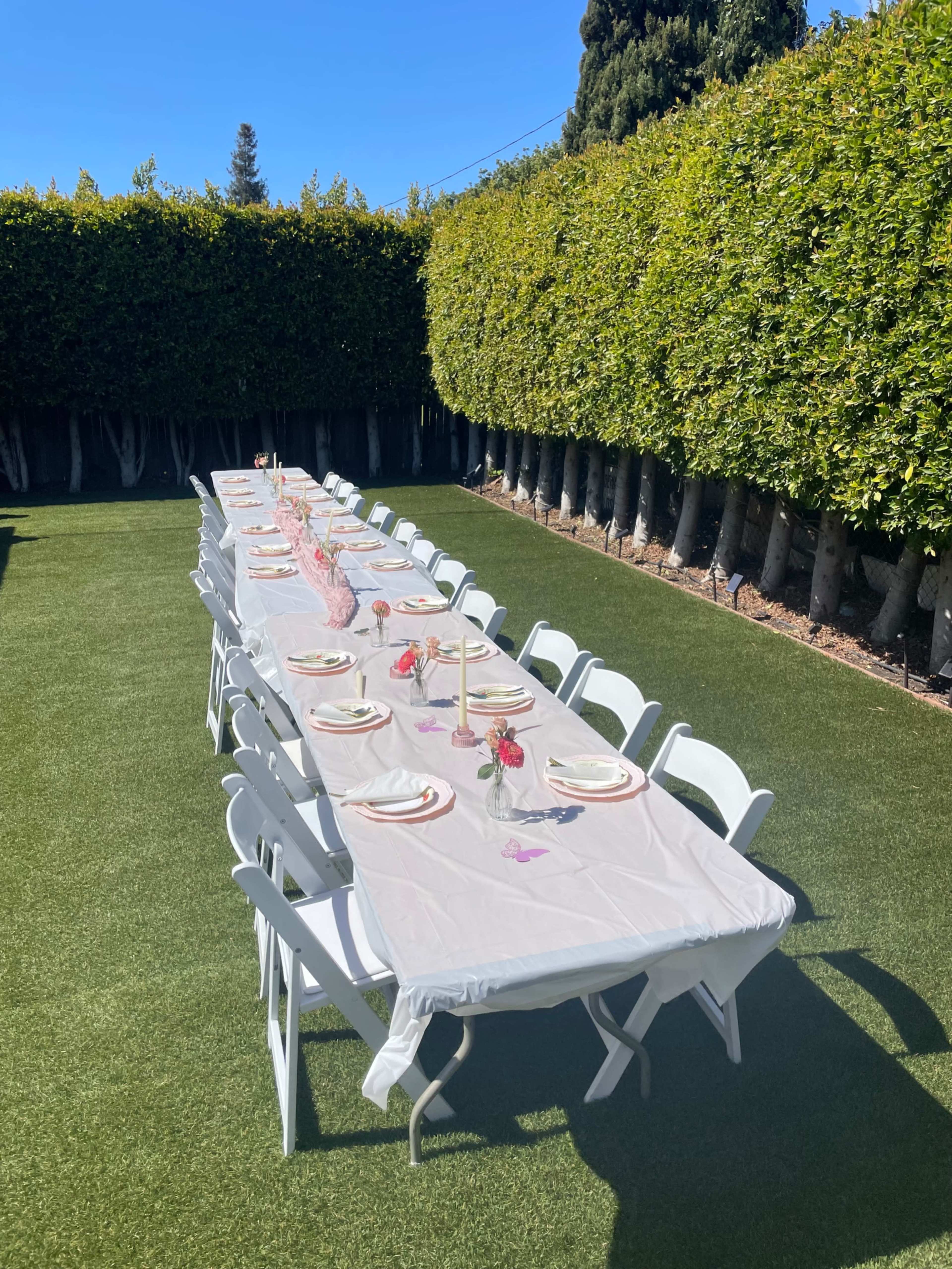 A long dining table is set outdoors on a grassy area, surrounded by tall hedges and accented with pink decorations.