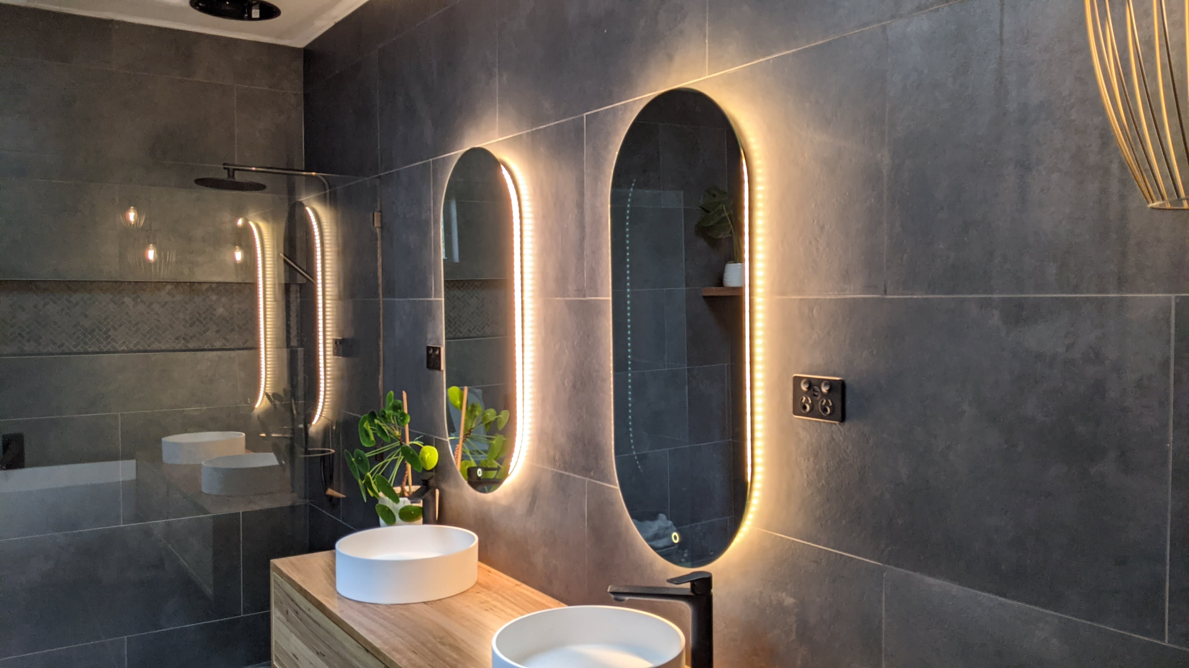 The image shows a modern bathroom with three oval mirrors featuring backlighting, a wooden vanity with two white vessel sinks, and wall-mounted fixtures.