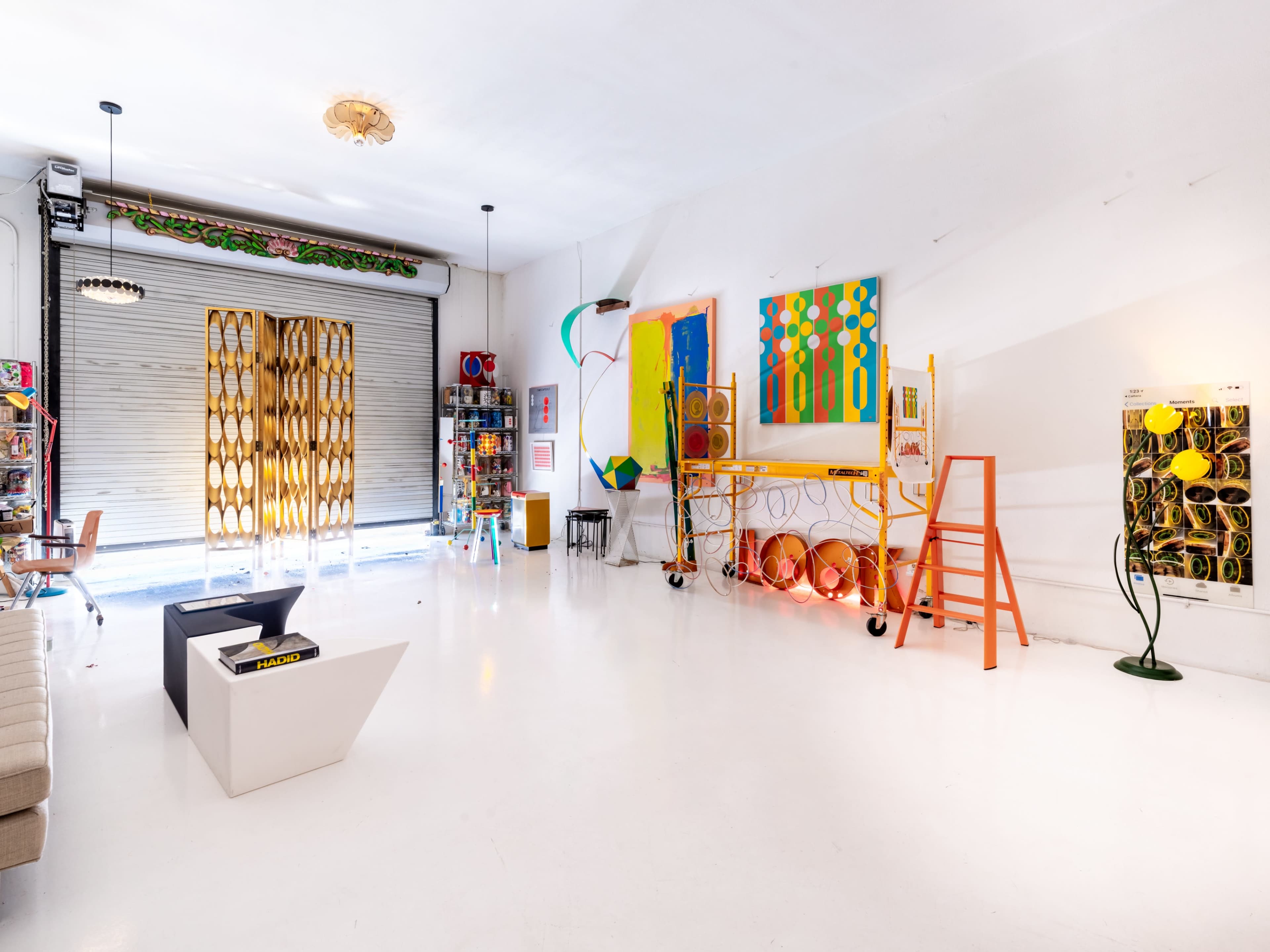 The image shows a brightly lit art studio featuring colorful paintings, various art supplies, and a sleek white floor.