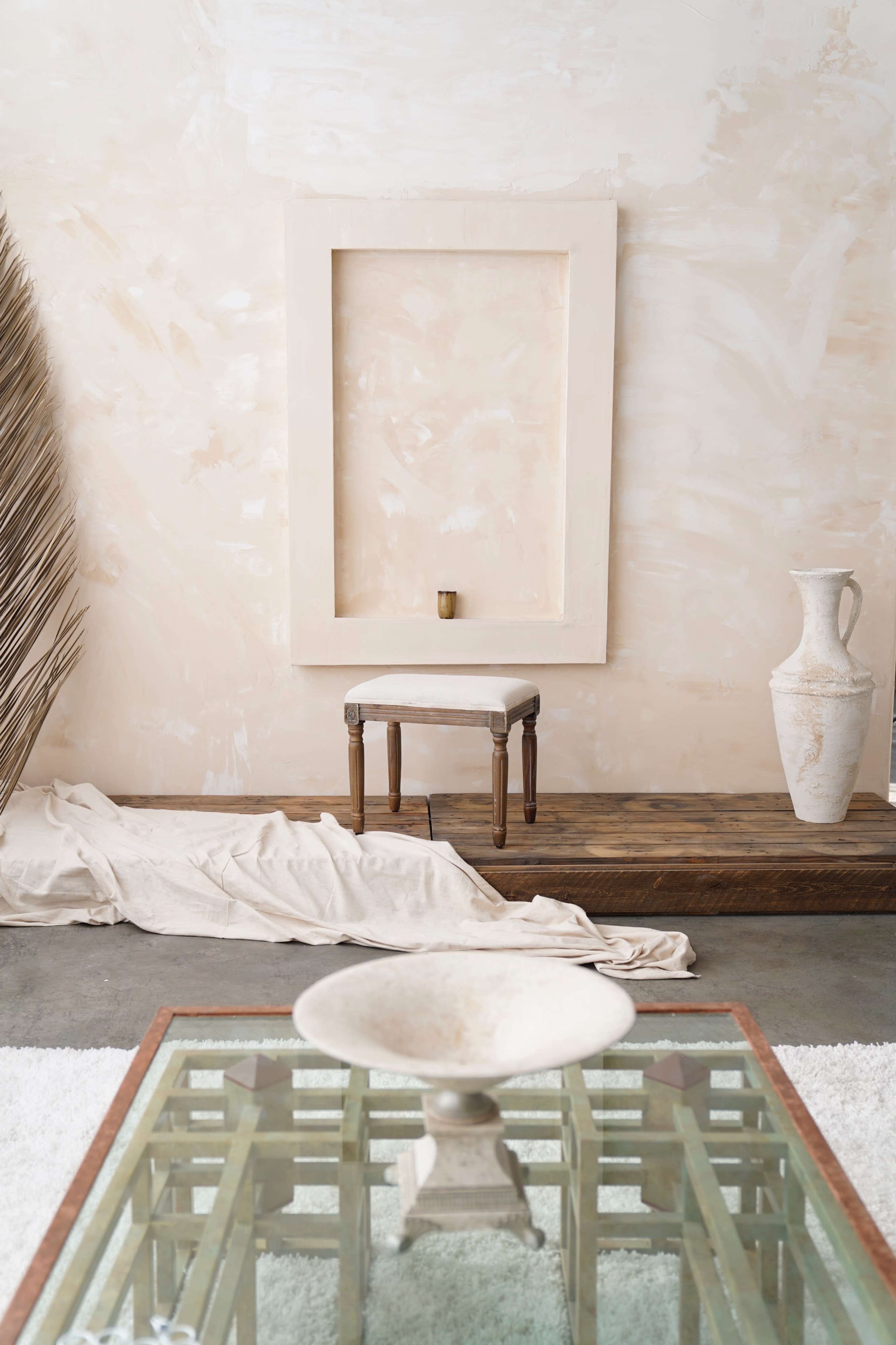 The image shows a minimalist interior space featuring a blank frame on the wall, a small stool, a wooden platform, a decorative vase, and a glass table with a shallow basin on top.