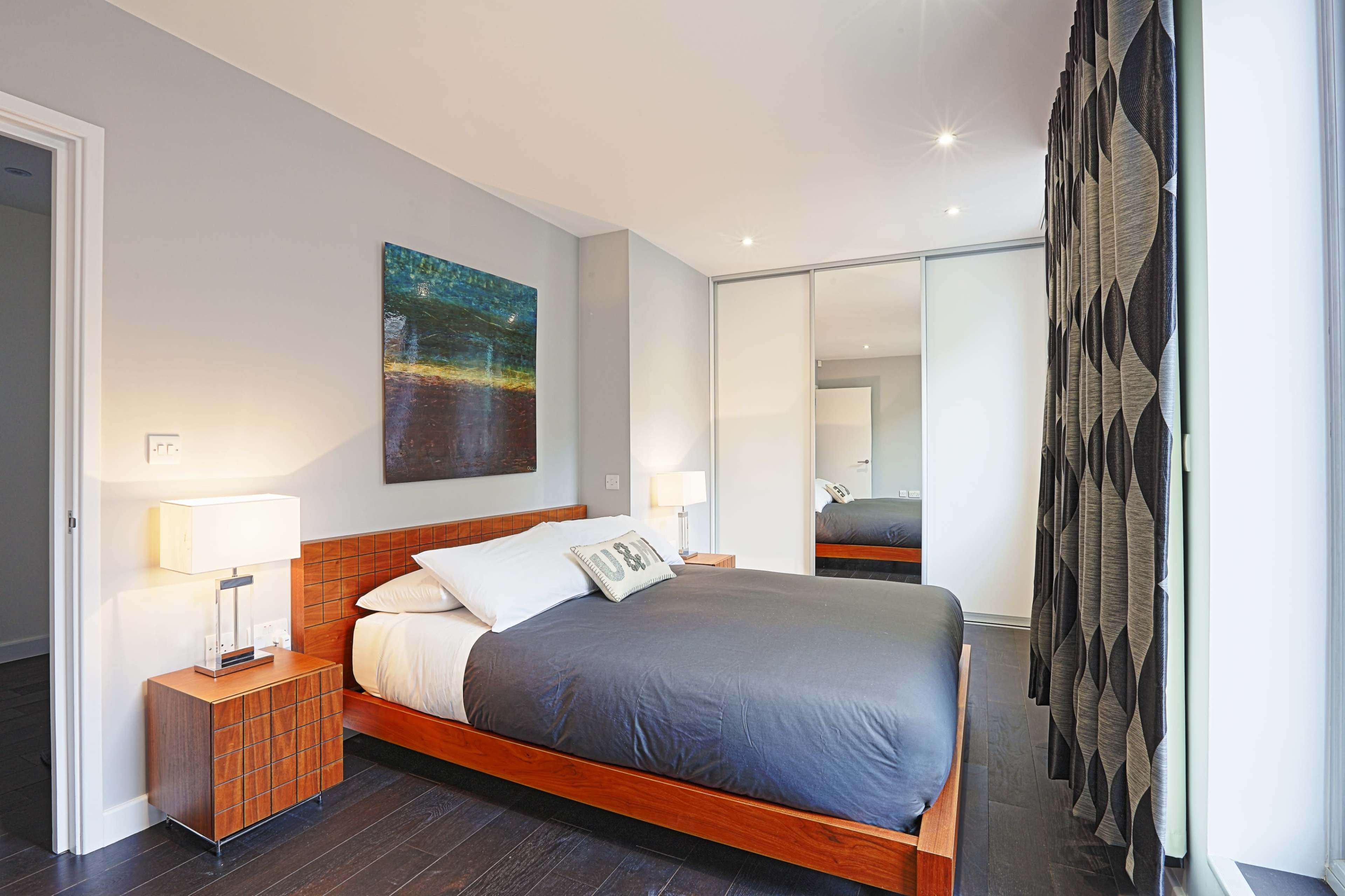 A modern bedroom with a wooden bed, two bedside lamps, a large mirror, and artwork on the wall.