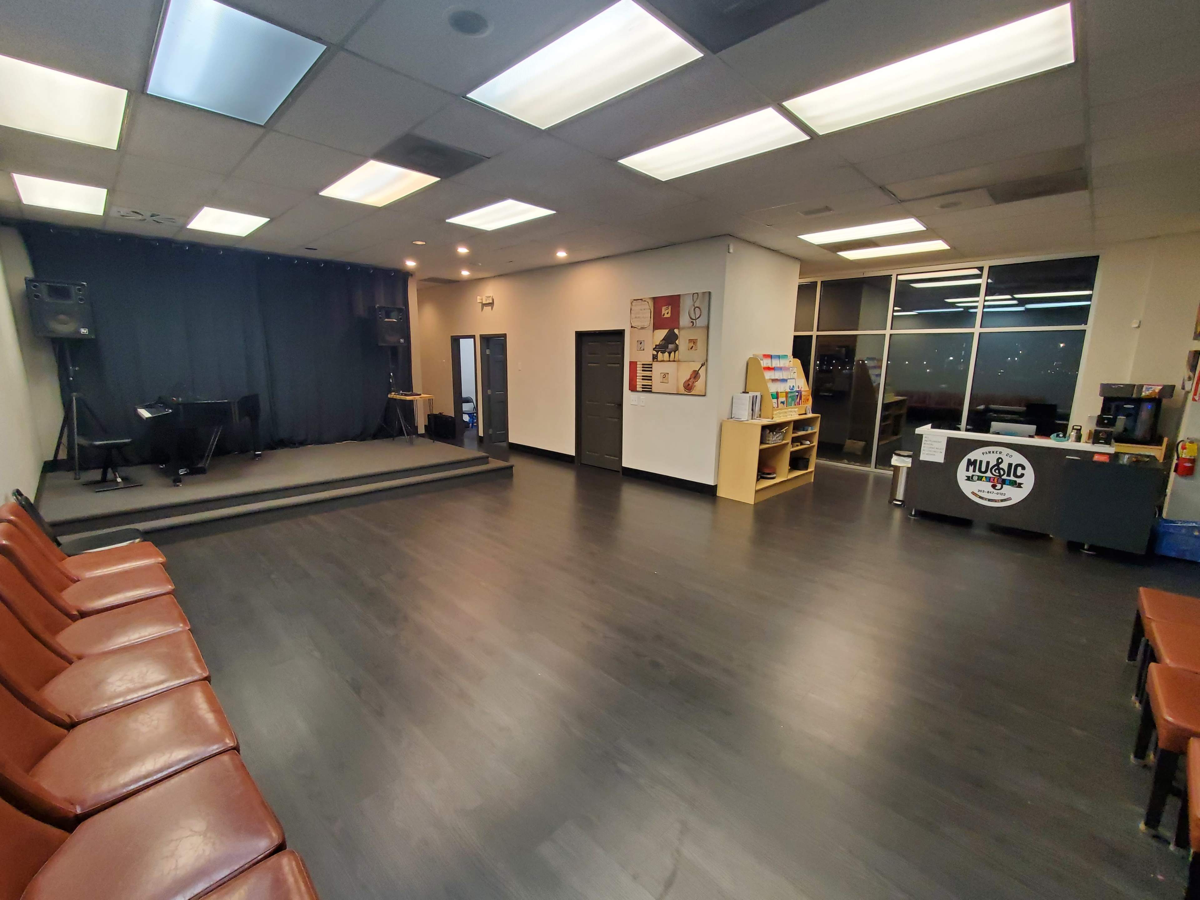 The image shows an empty music studio with a black stage at the front, seating along one side, and a refreshments area on the right.