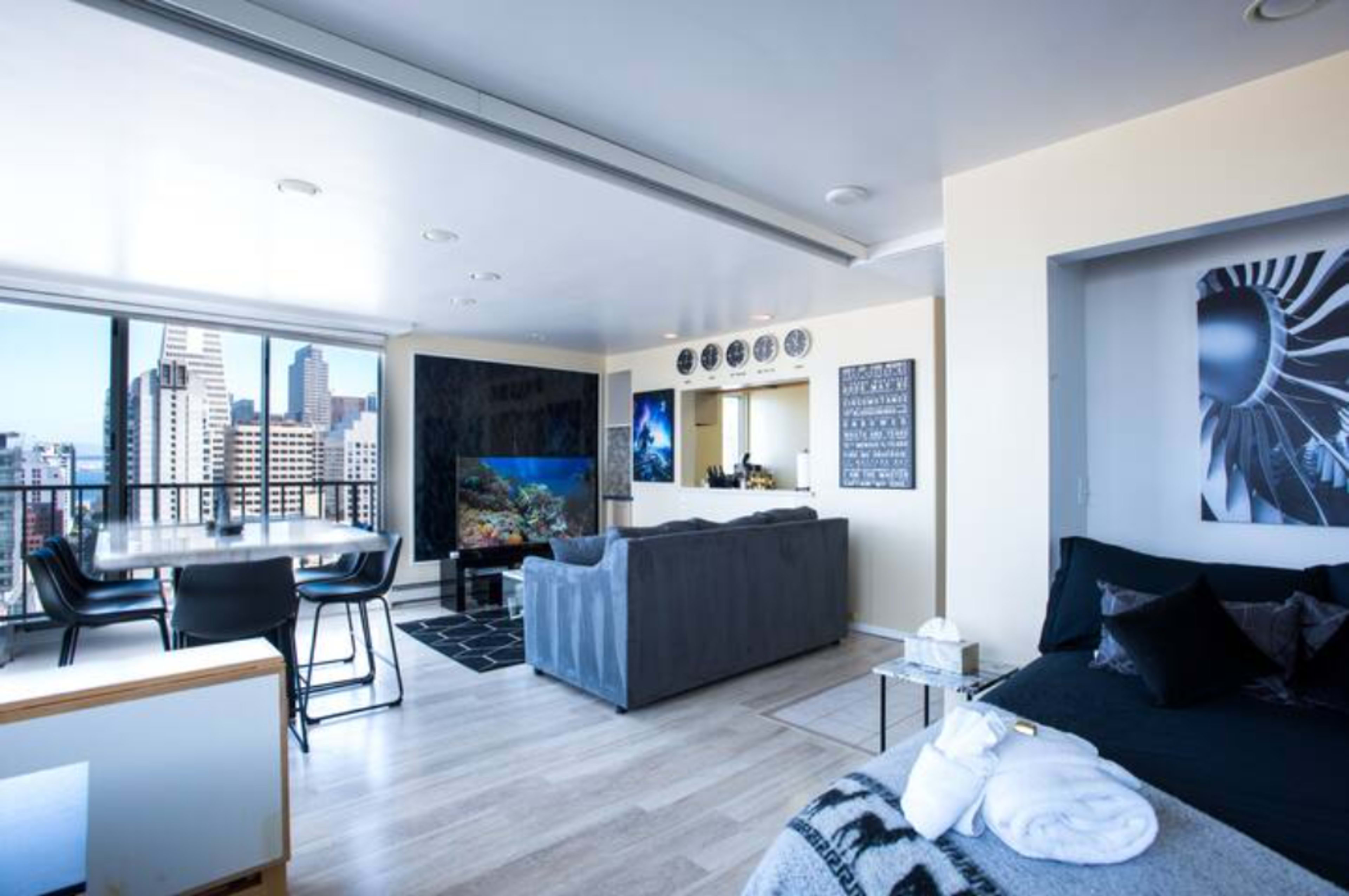 The scene shows a modern apartment interior with a living area, dining space, kitchen, and large windows offering a view of city buildings.