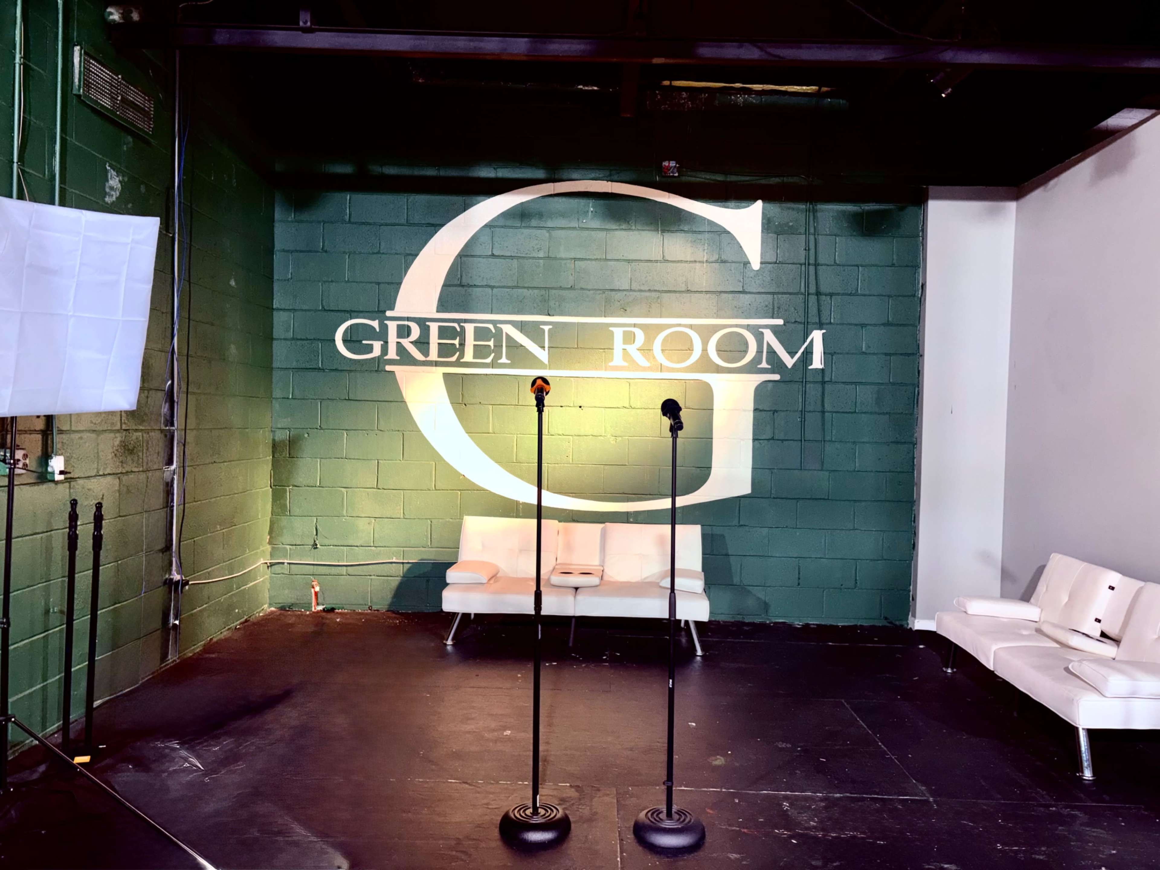 The image shows a green room with a large "GREEN ROOM" sign on the wall, two microphone stands in front, and a white seating area.