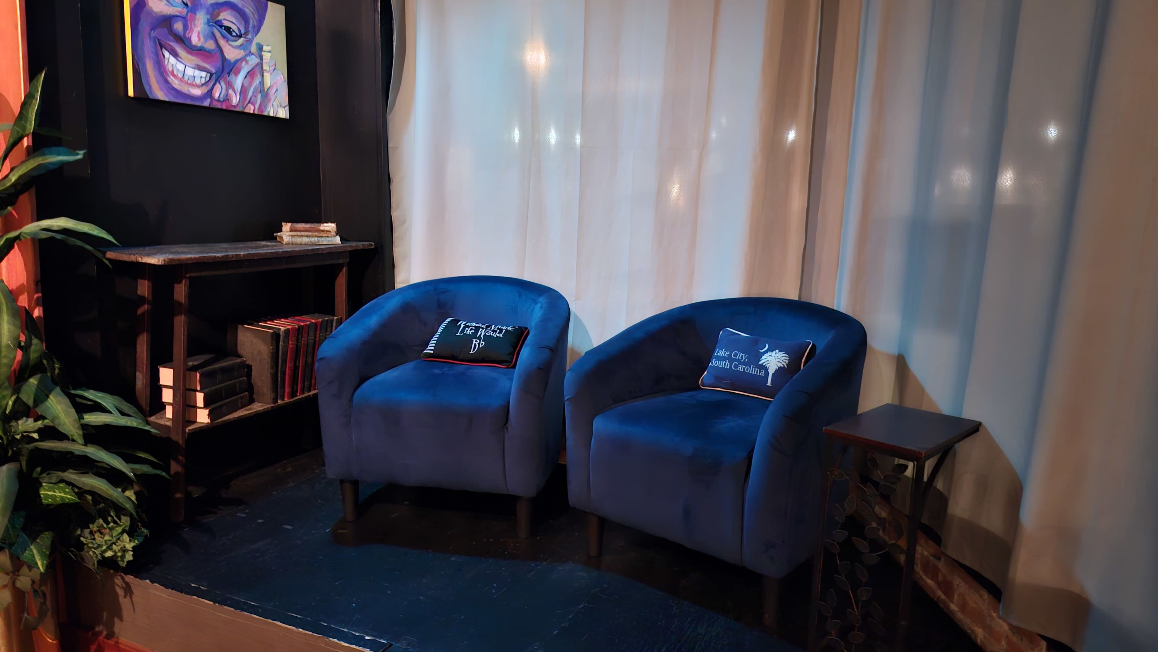 The image shows two blue upholstered chairs with decorative pillows positioned near a dark wooden shelf against a backdrop of white curtains.
