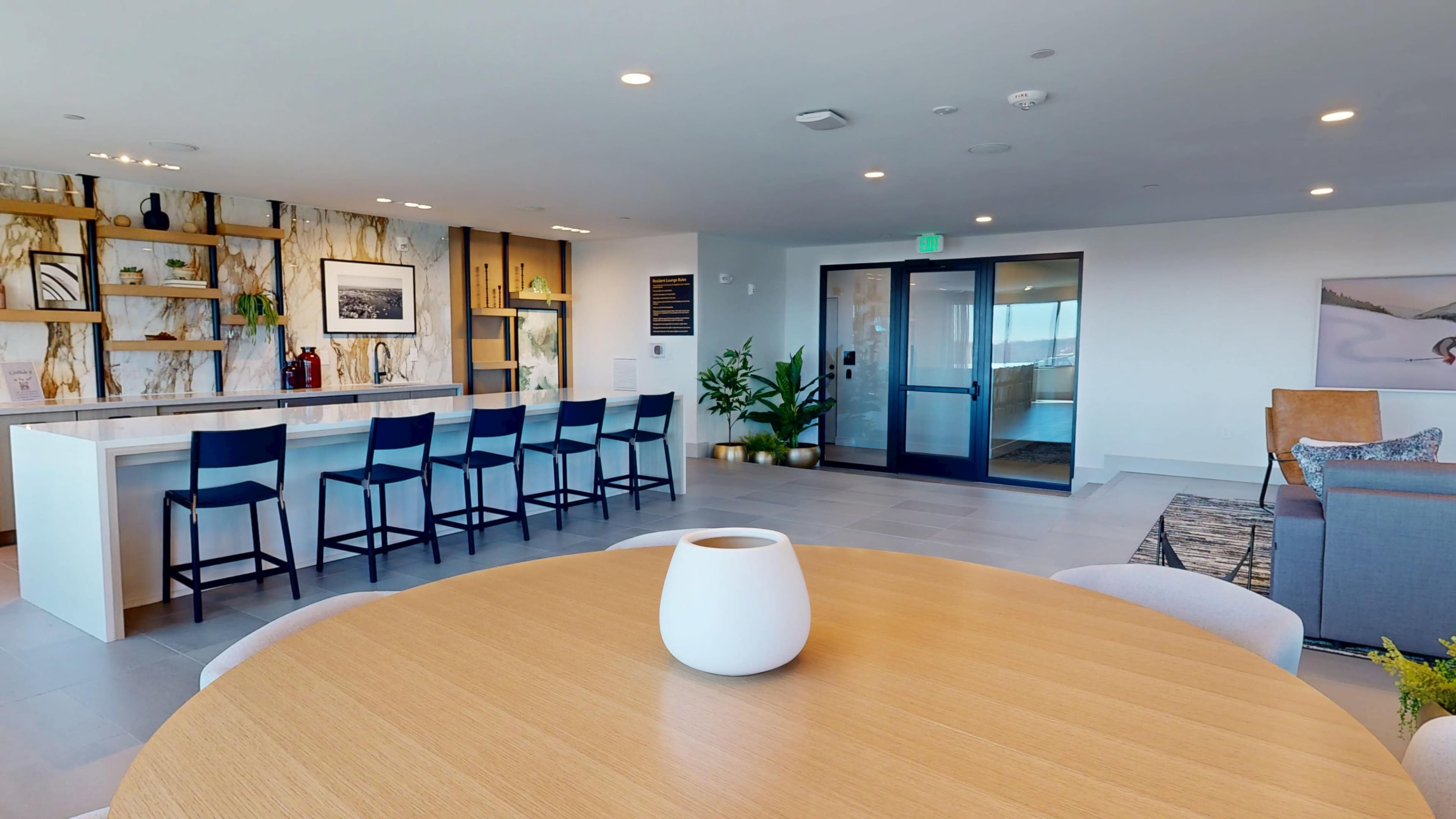 The image shows a modern lounge area featuring a circular wooden table, a bar with several stools, and large windows leading to an exterior view.