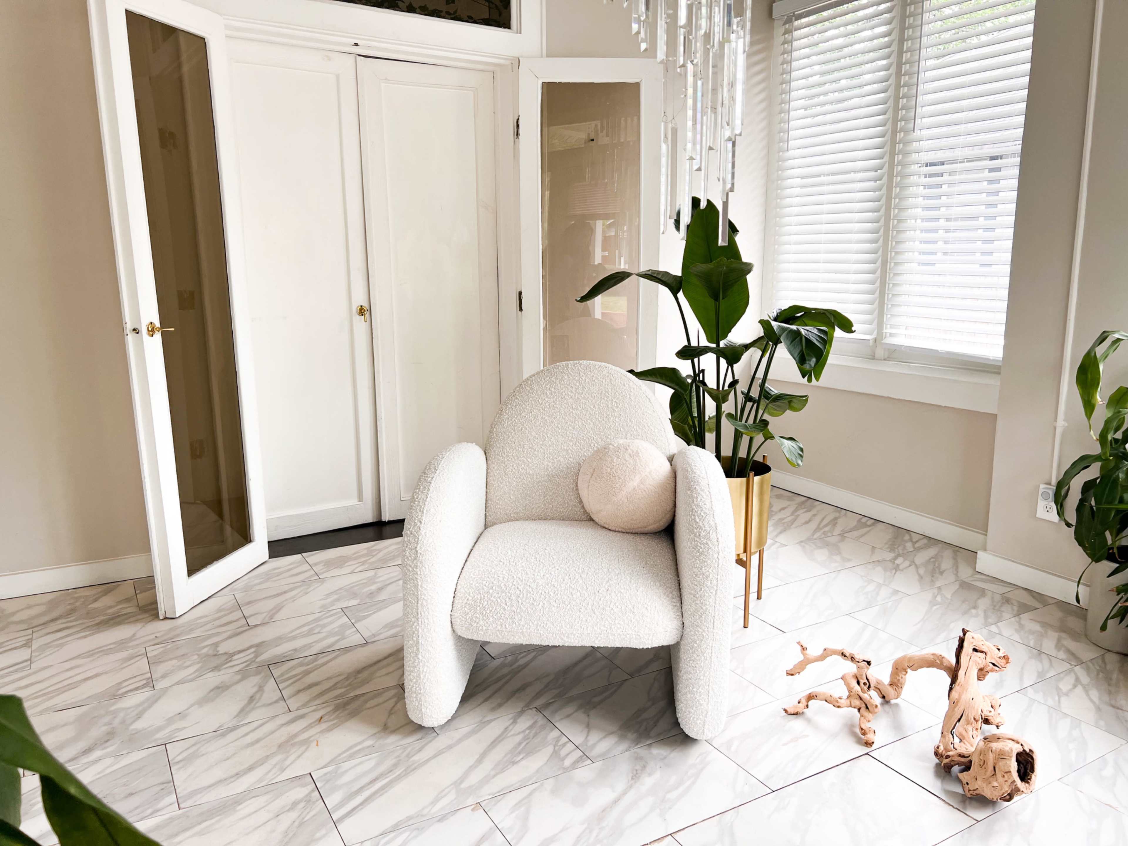 The image features a textured white armchair positioned beside a potted plant in a well-lit room with tiled flooring and large windows.