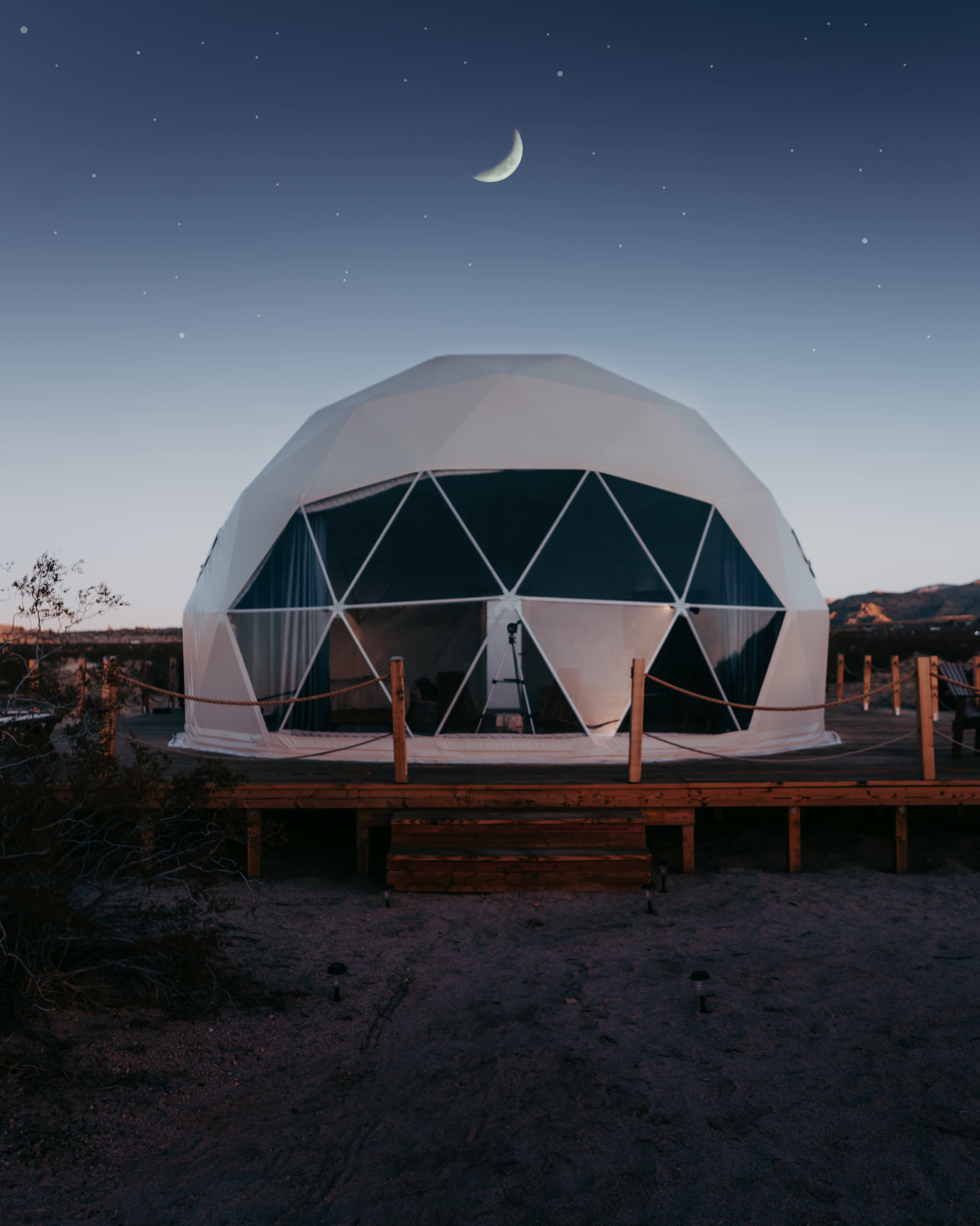 A geodesic dome with large triangular windows is situated on a wooden platform under a crescent moon and starry sky.