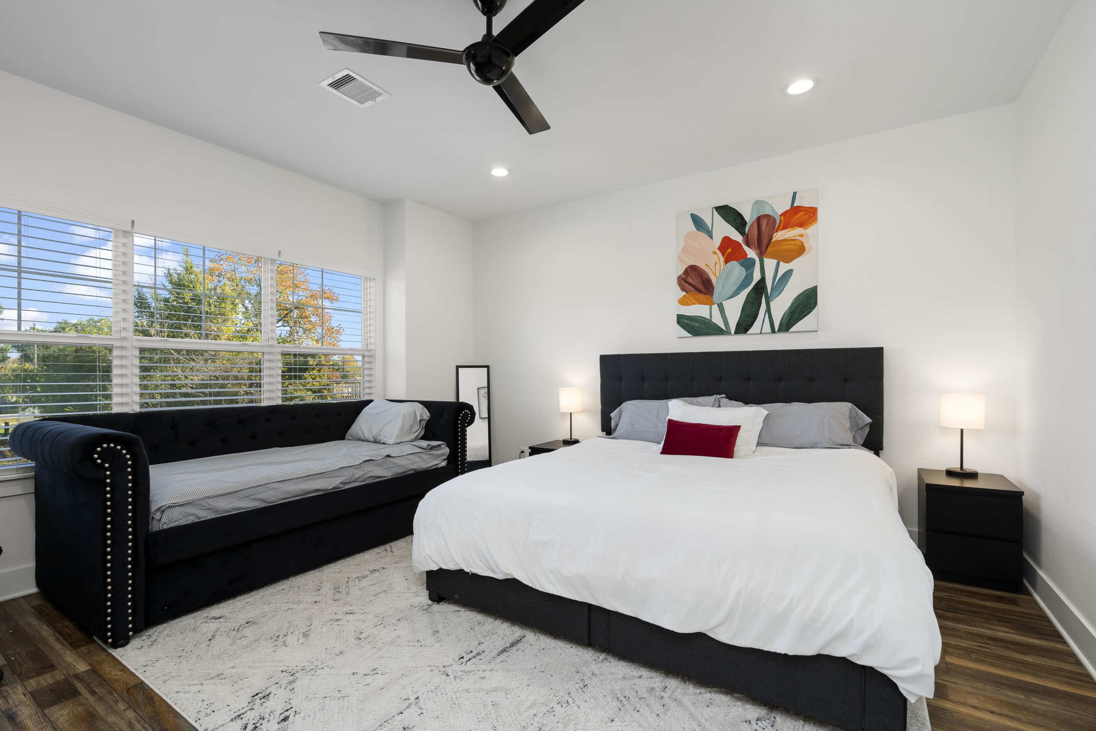 A modern bedroom featuring a queen-sized bed with white linens, a black sofa against one wall, and large windows with blinds allowing natural light to enter.
