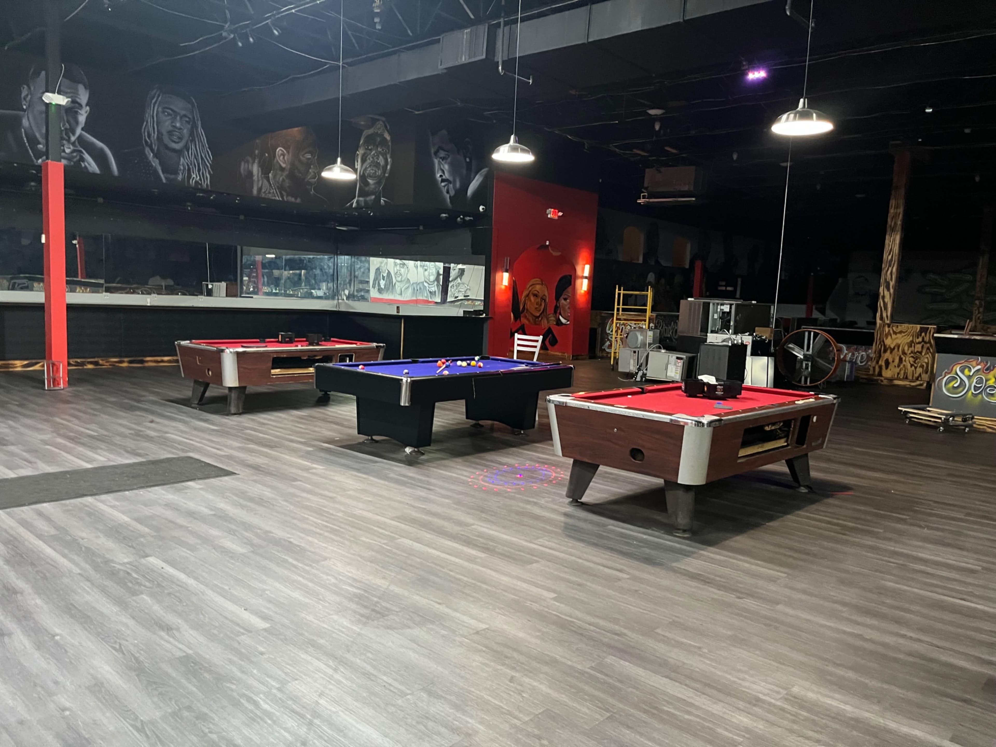 The image shows an indoor recreational space with four pool tables arranged in a large area featuring wall murals and dim lighting.