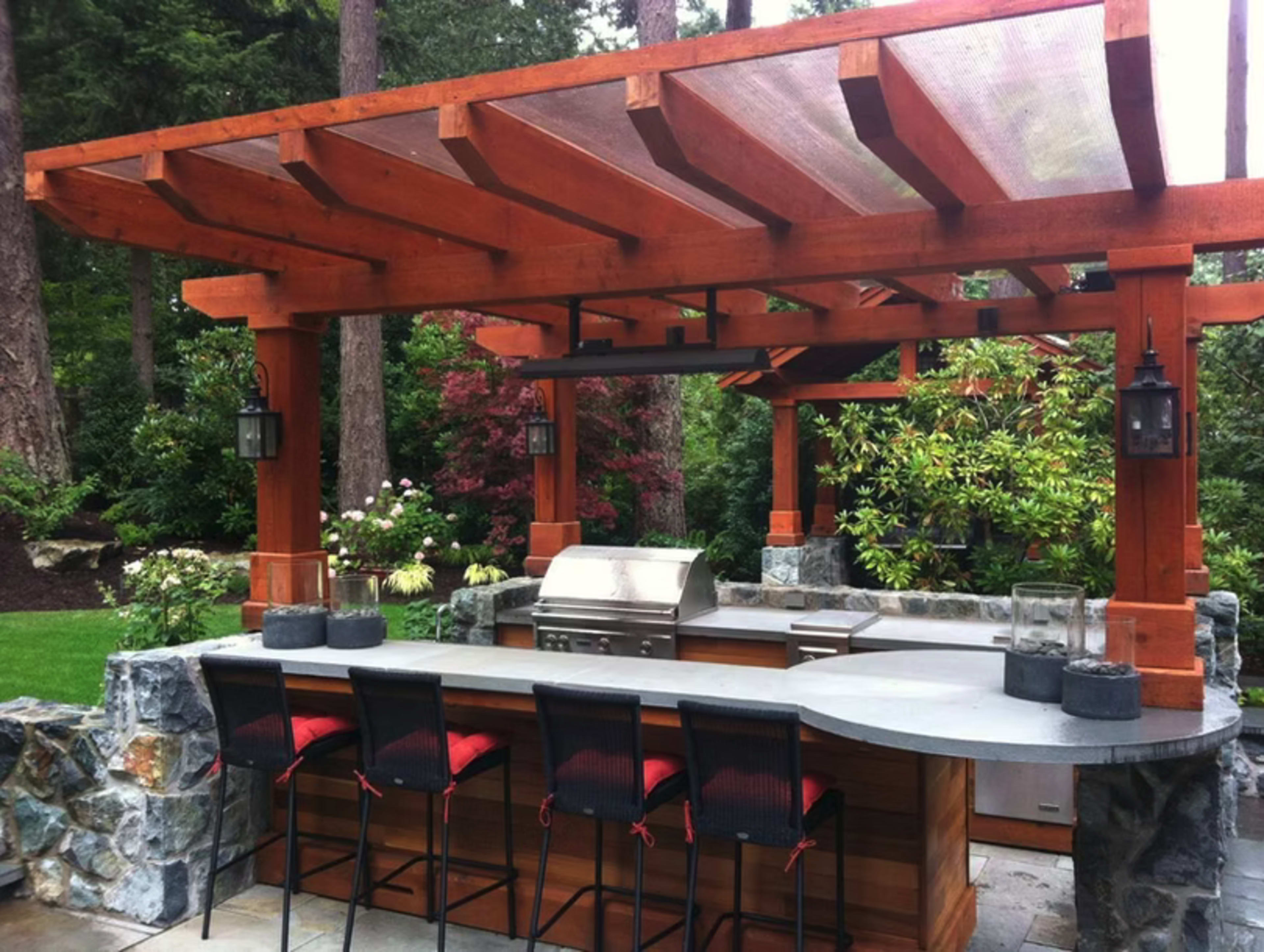 The image shows an outdoor kitchen with a stone bar area, a grill, and wooden pergola, surrounded by lush greenery.