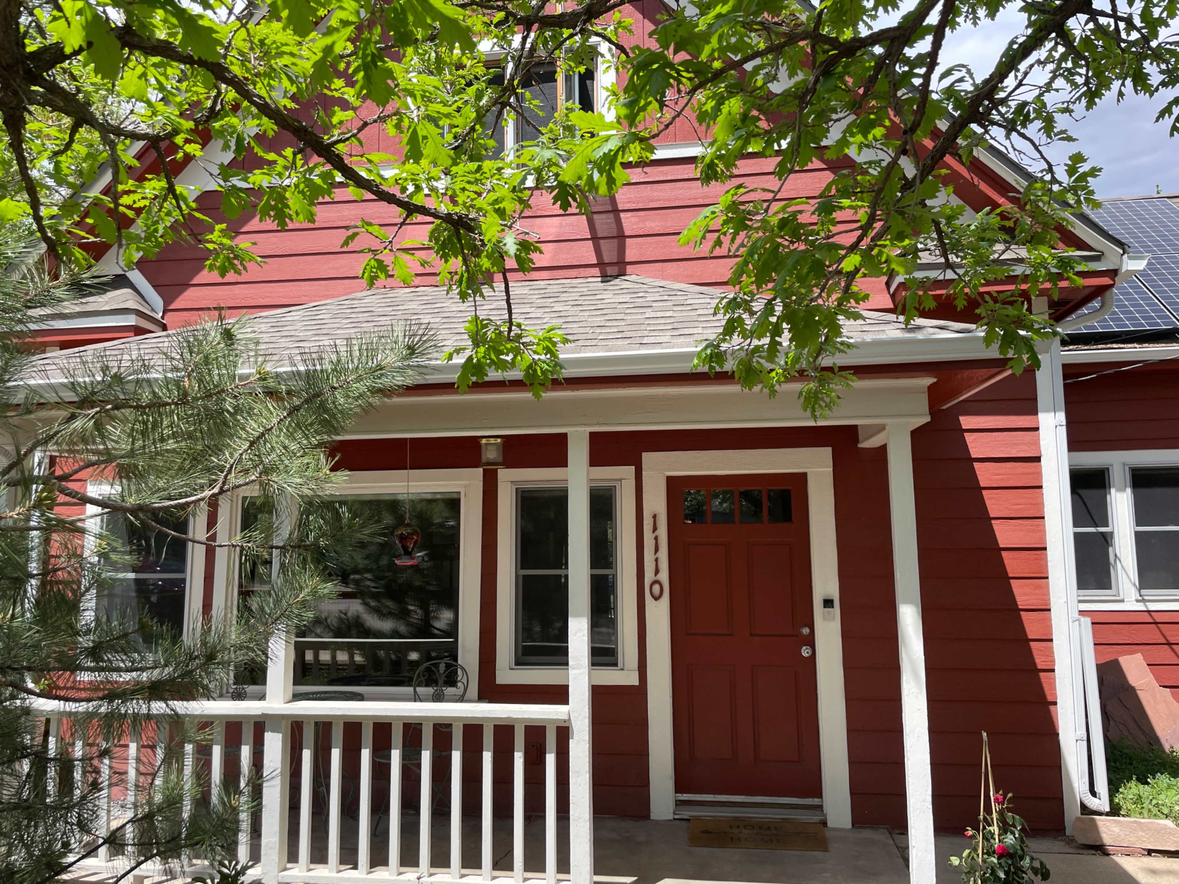 A red two-story house with a sloped roof and a front porch is surrounded by green leaves and trees.