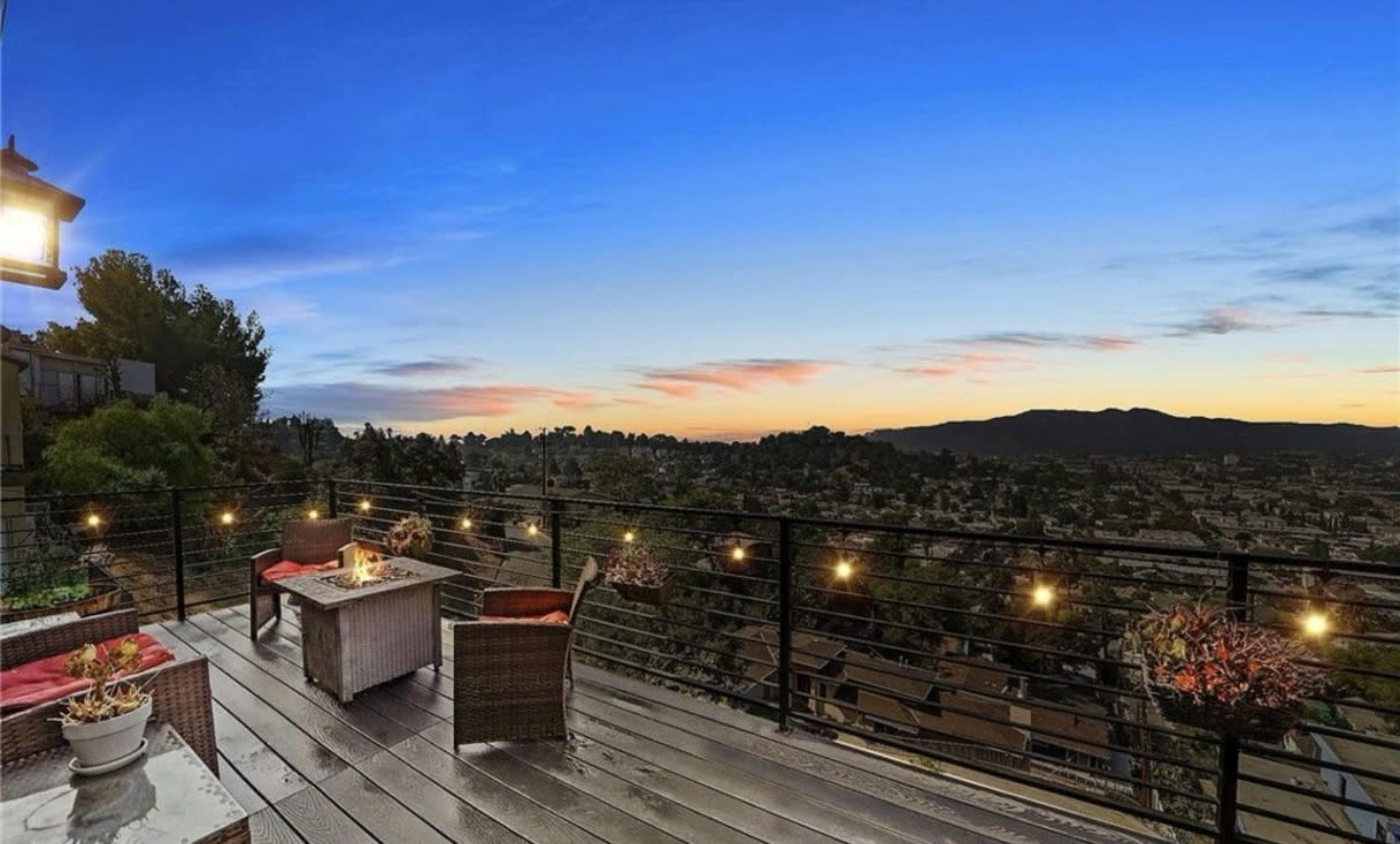 The image shows a rooftop patio with outdoor furniture overlooking a scenic view of a city and mountains at twilight.