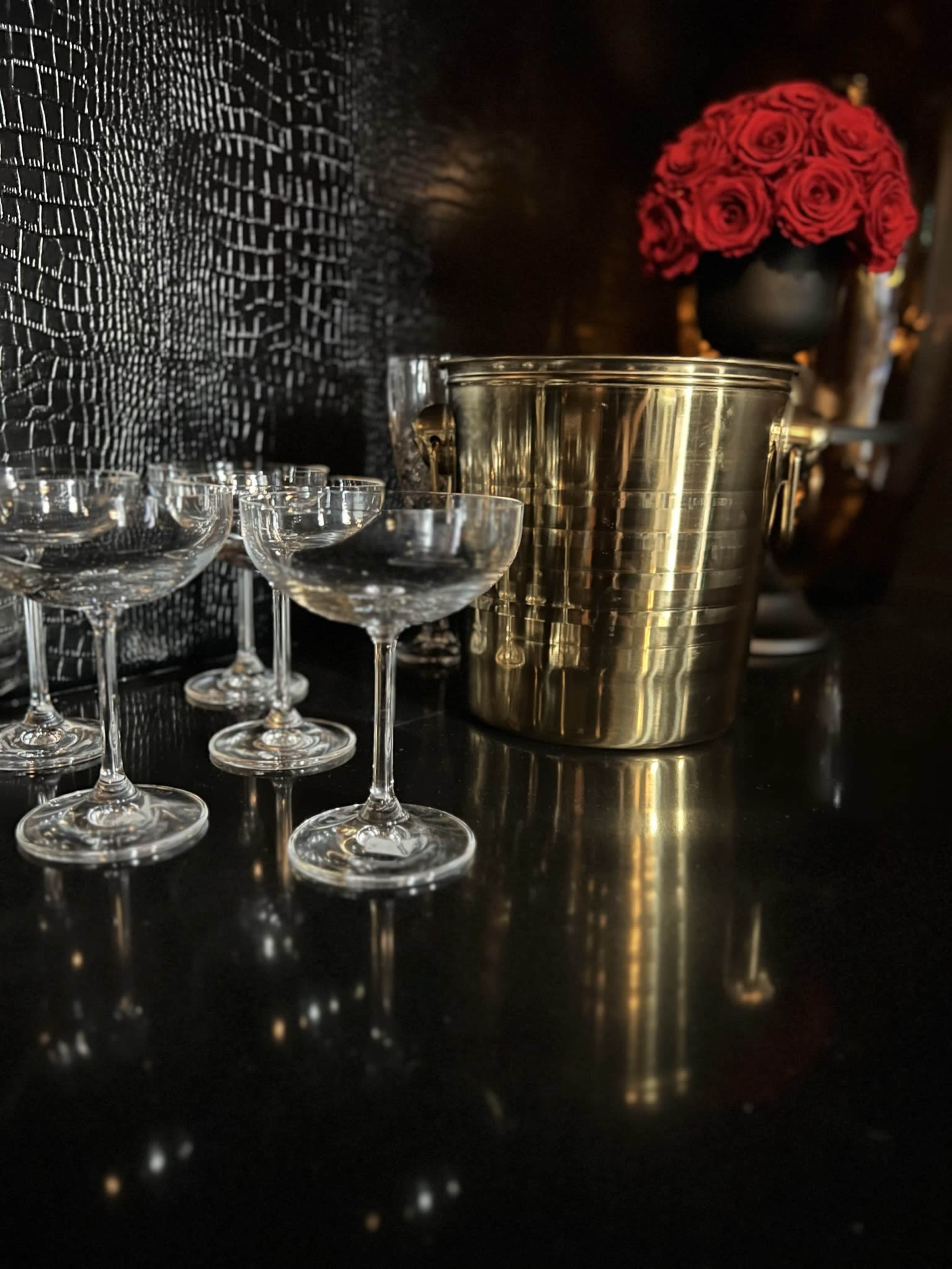 A polished brass ice bucket sits beside several crystal champagne coupes and a vase of red roses on a black countertop.