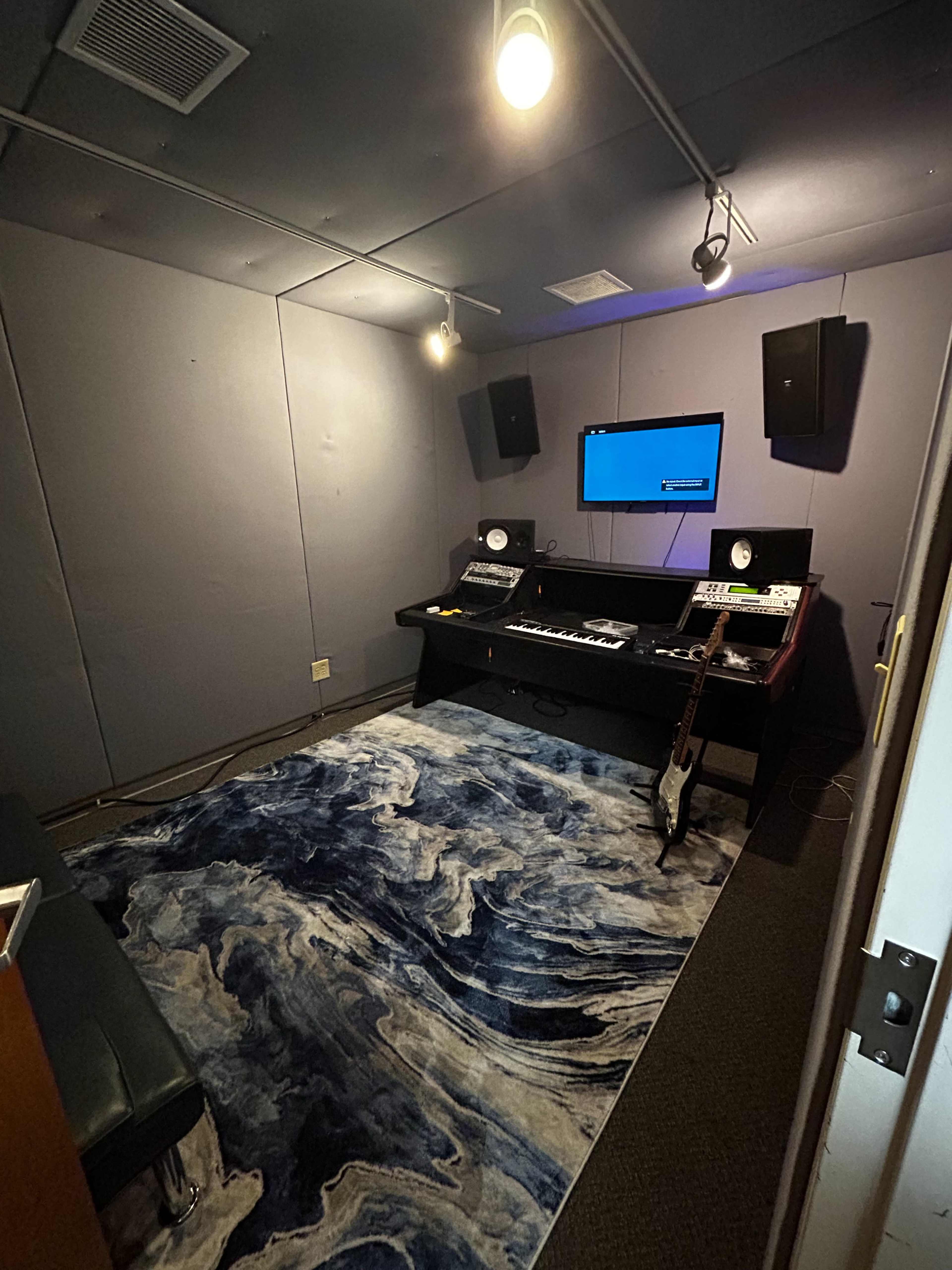 A small music studio with a soundproofed interior, featuring a desk with audio equipment, a large screen, and a rug on the floor.