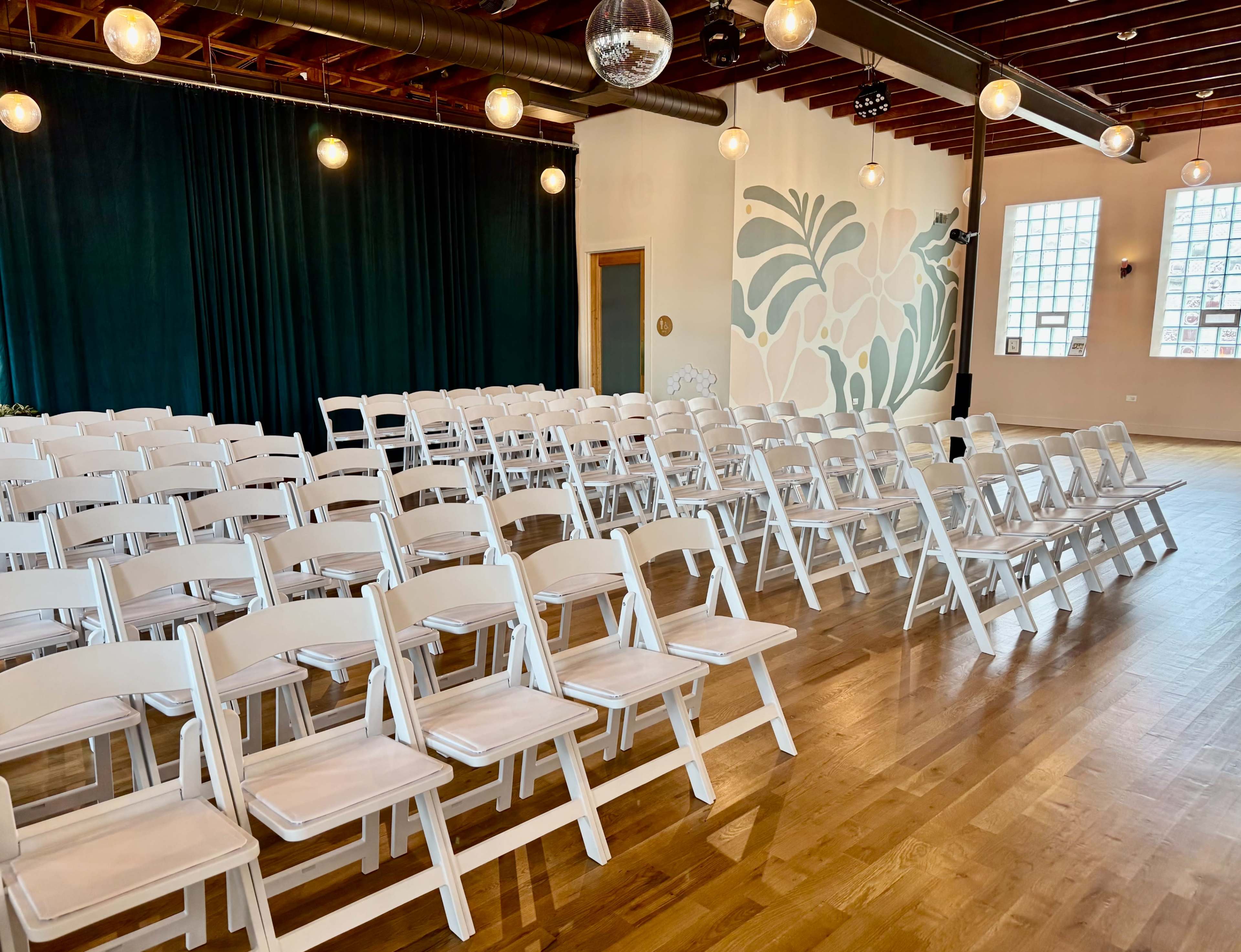 The image shows a spacious room set up with rows of white folding chairs facing a green curtain and a decorated wall with a mural.