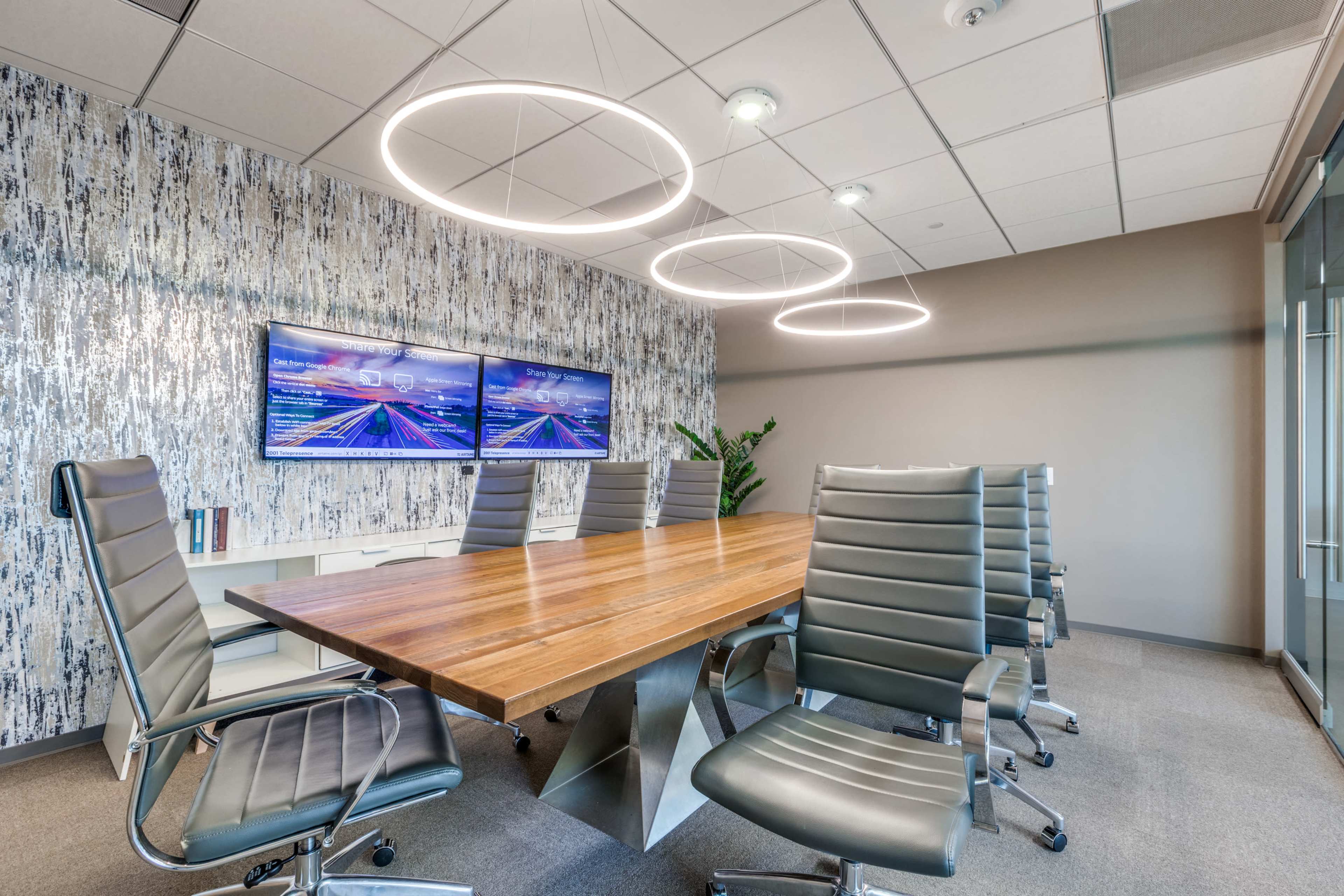 The image shows a modern conference room featuring a large wooden table, six ergonomic chairs, and two wall-mounted screens, all illuminated by circular overhead lights.