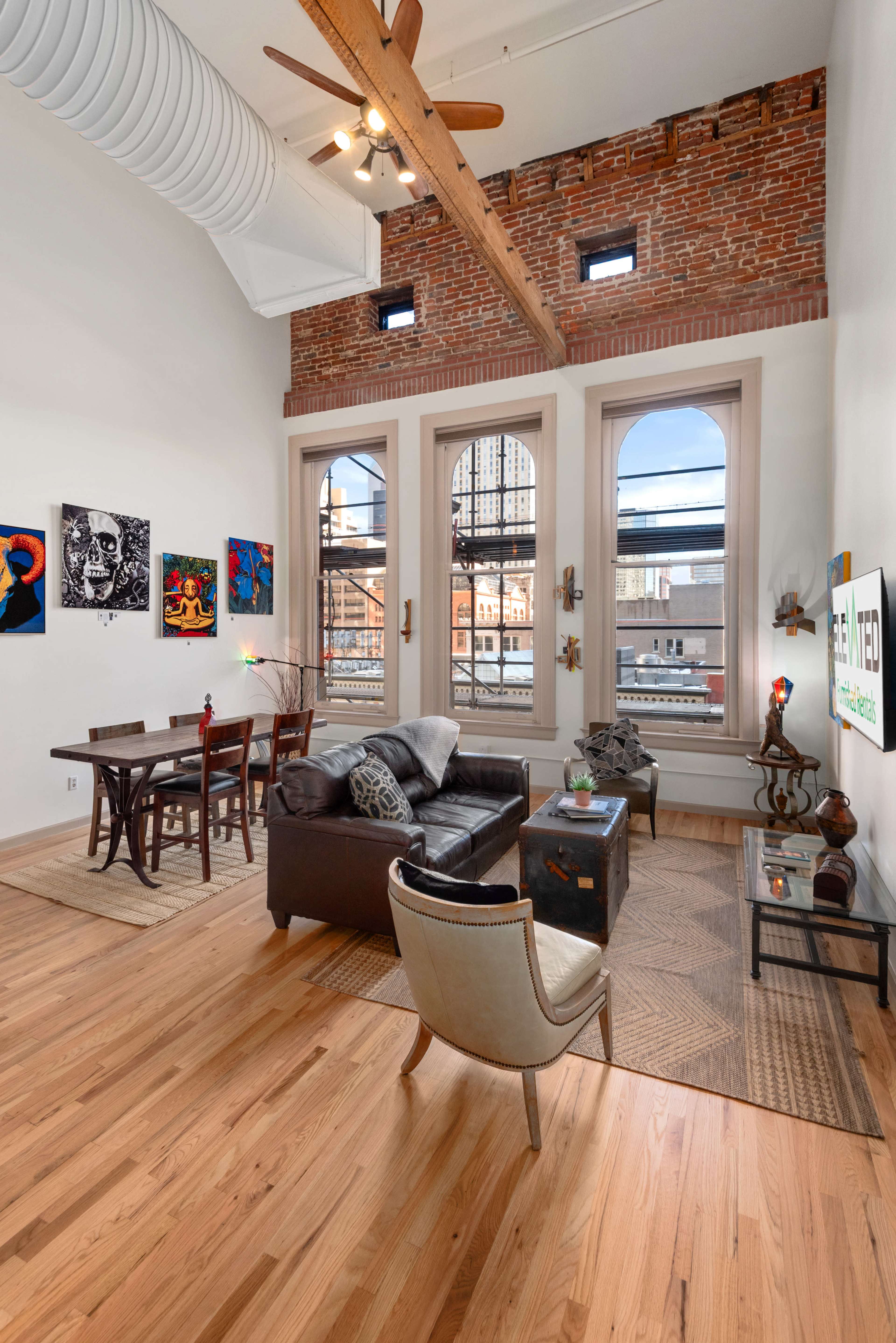 The image shows a spacious living area with a brick wall, large windows, and wooden floors, furnished with a dark leather sofa, a dining table, and various artworks on the walls.
