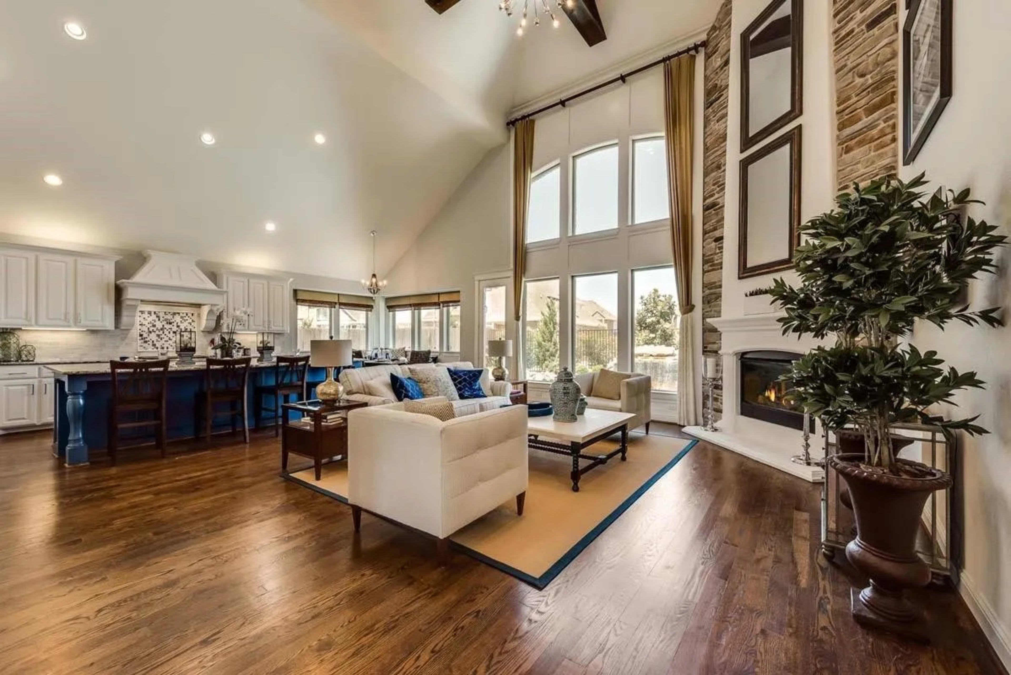 The image shows a spacious living area featuring a white sofa and a coffee table, with a stone fireplace and large windows that let in natural light.