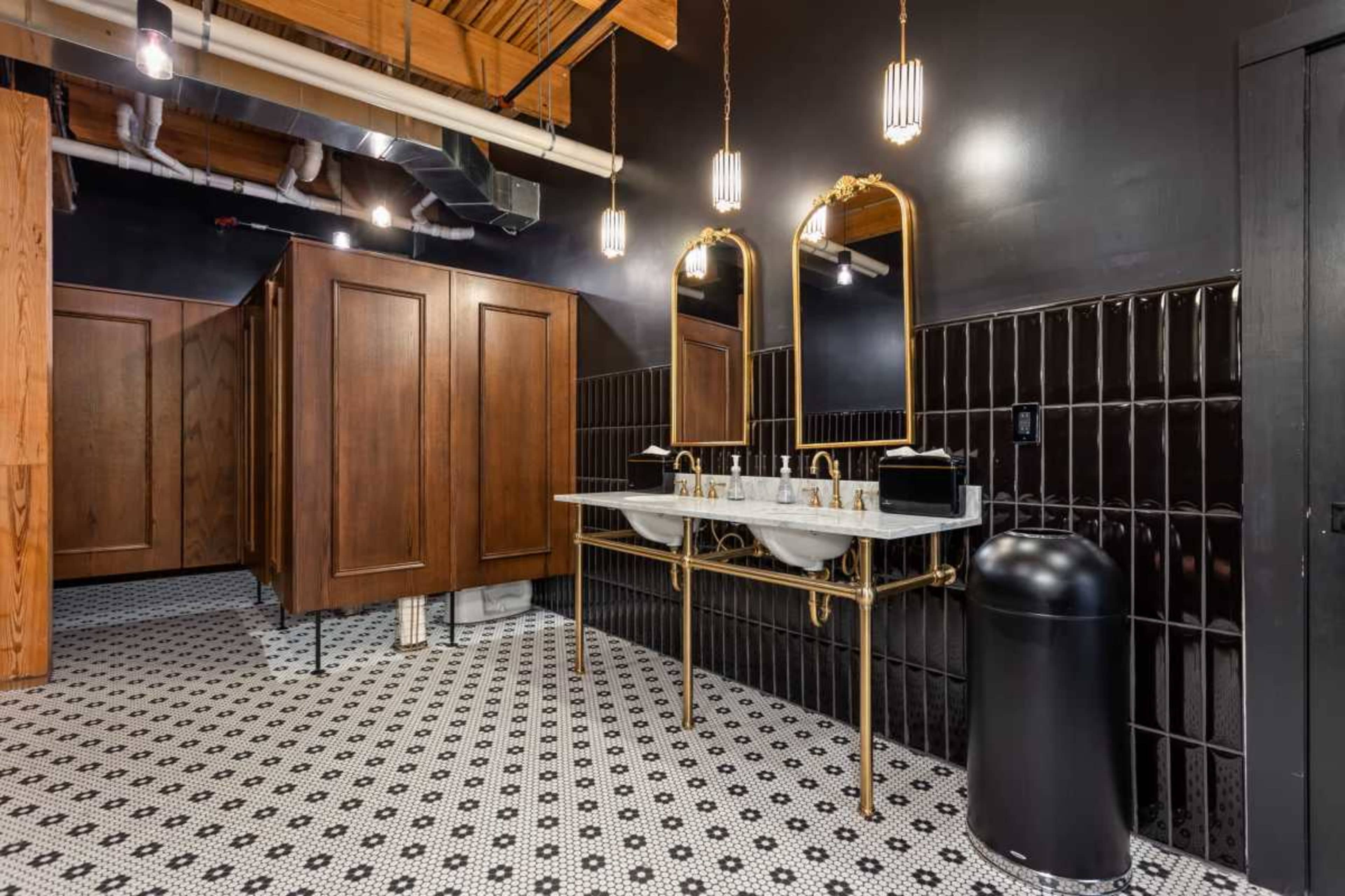 The image shows a modern bathroom with black walls, two stylish sinks on a marble countertop, and wooden stalls in the background.