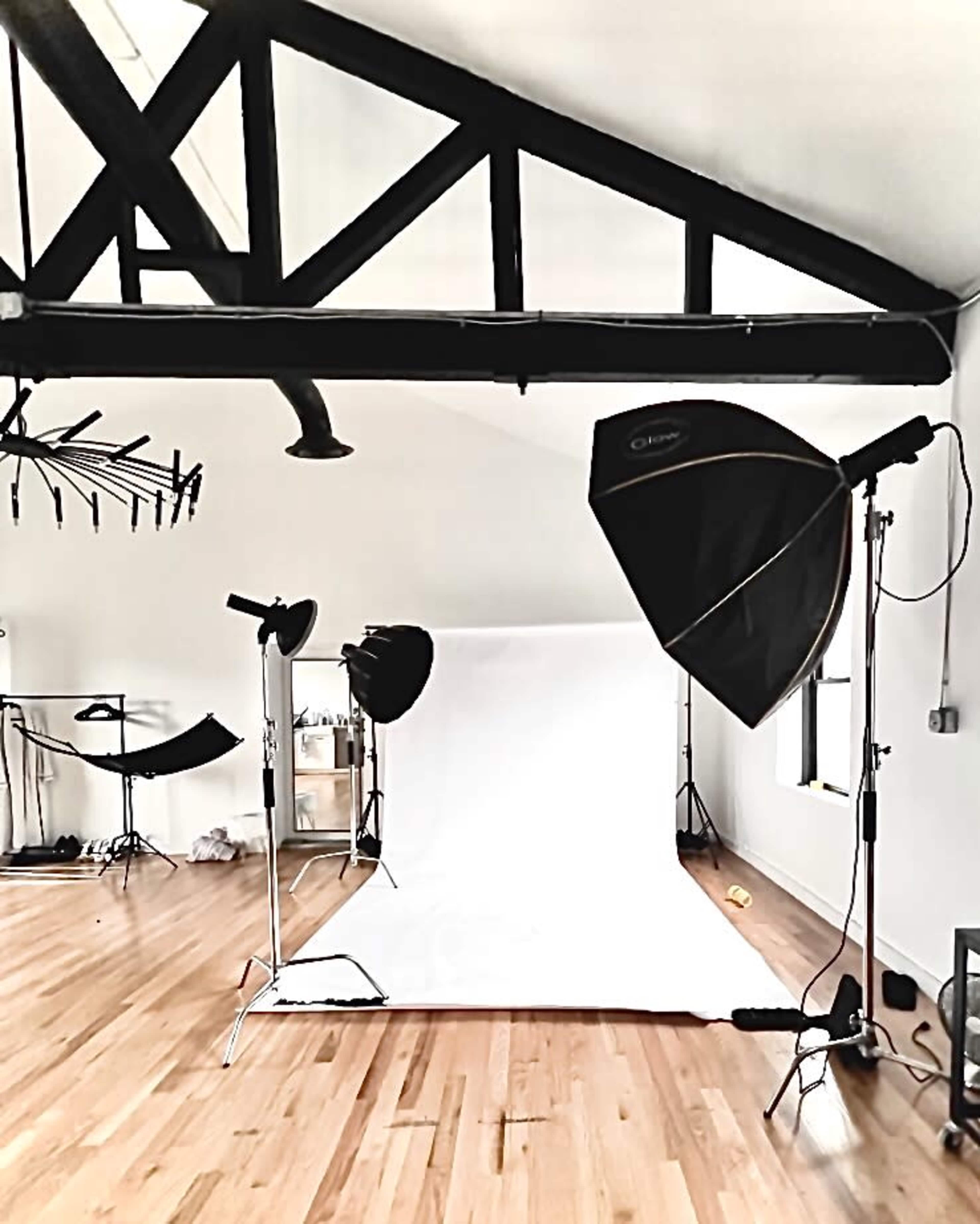 The image depicts a photography studio with a white backdrop, several lighting equipment setups, and wooden flooring.