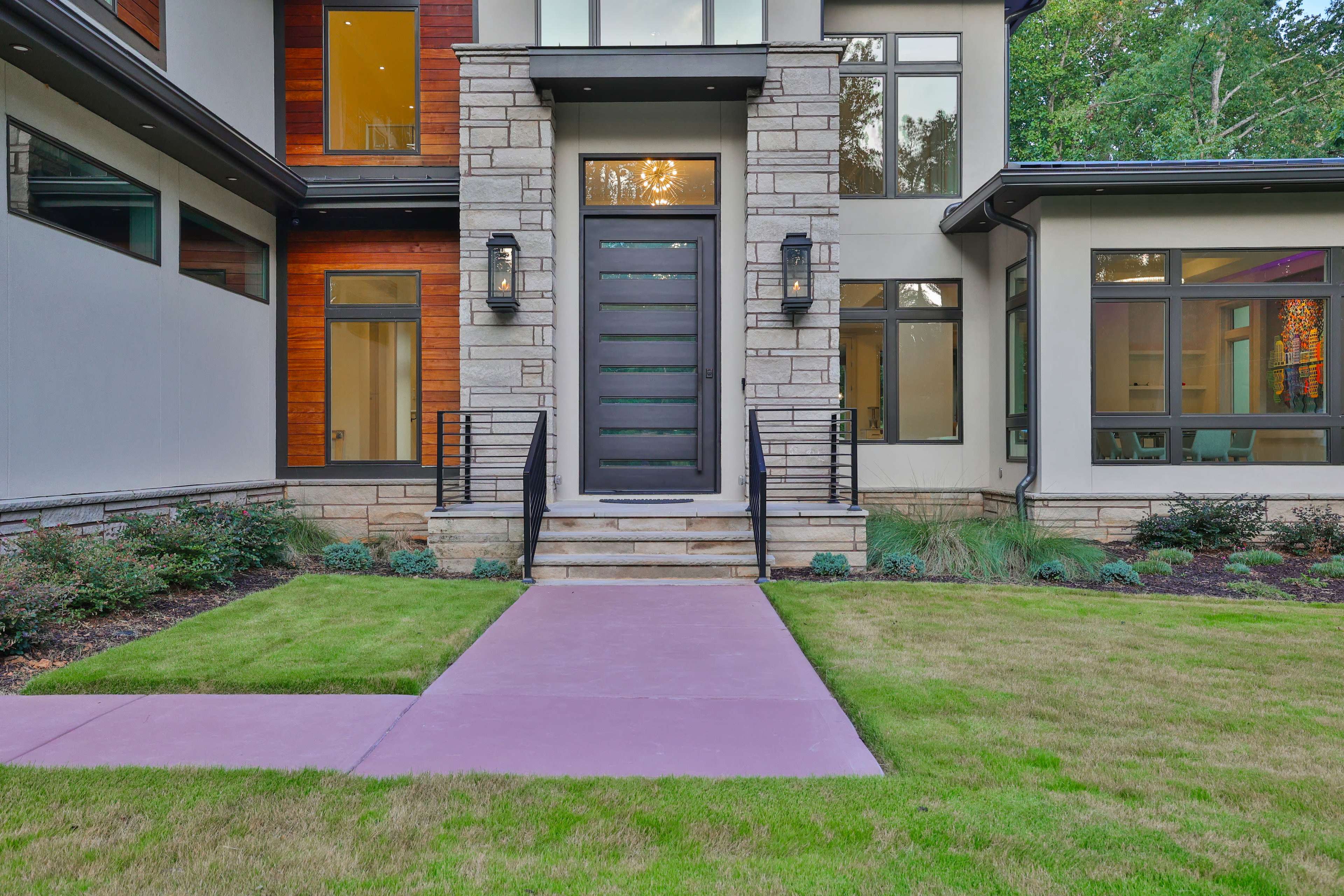 The entrance of a modern home featuring a large black front door, stone accents, and a landscaped lawn with a purple walkway leading to the door.