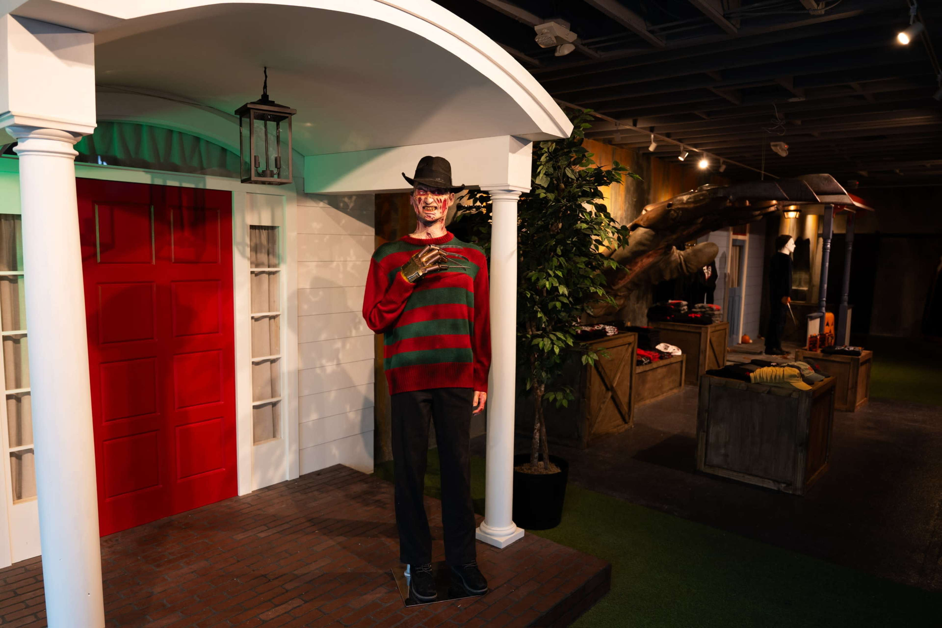 A life-sized figure dressed as a horror character stands on a porch with a red door, surrounded by a themed indoor setting.