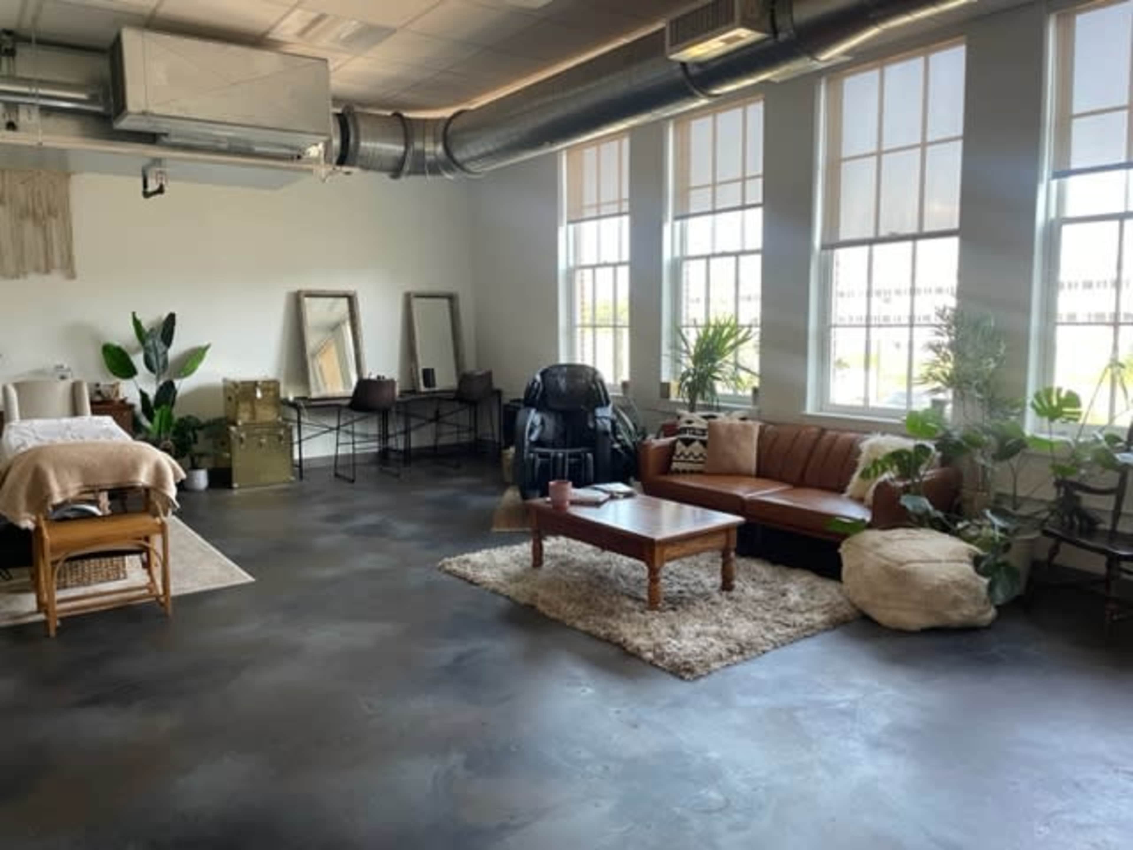 A spacious, well-lit room with large windows, featuring a mix of furniture including a brown sofa, a wooden coffee table, and a bed with textile elements, alongside several plants and mirrors.