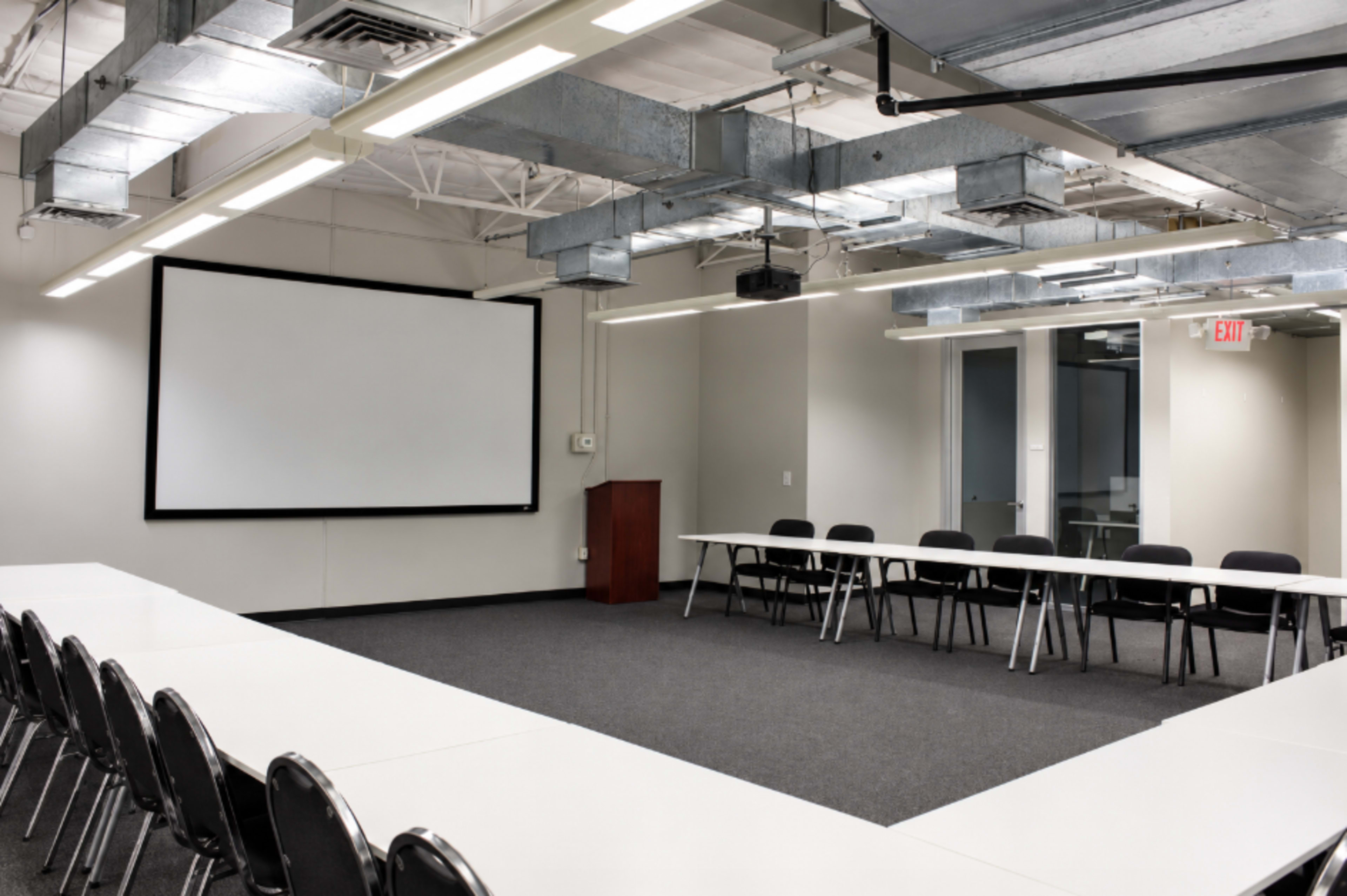 A spacious conference room features a large screen and two rows of tables arranged in a U-shape, with black chairs placed around them.