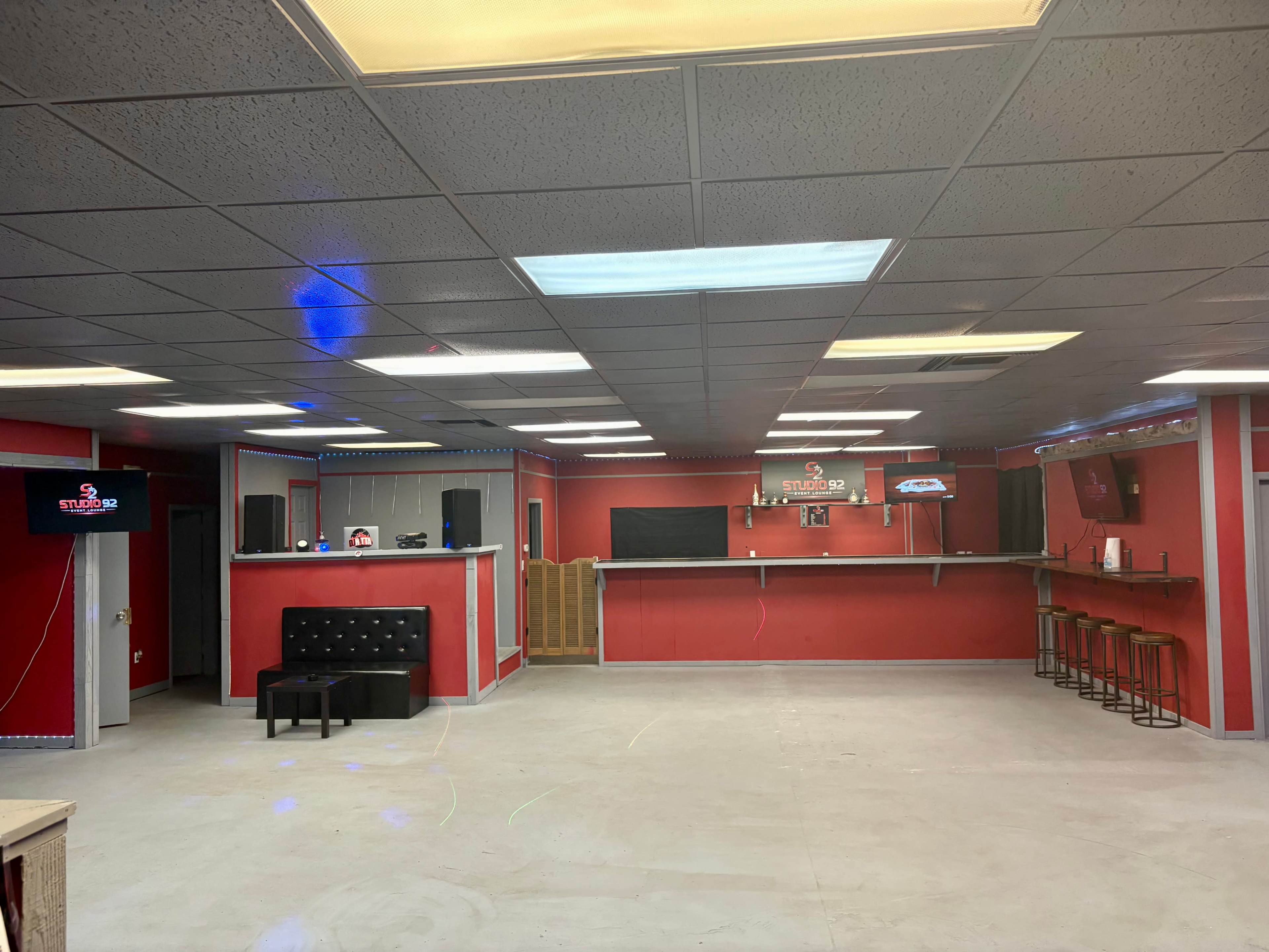 The image shows a spacious red room with a bar area, black seating, and multiple speakers on the walls.